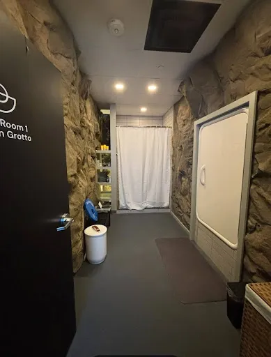 Interior of a room with stone-textured walls, a white curtain at the far end, a small shelf with items, a white trash bin, and a large wall-mounted panel.