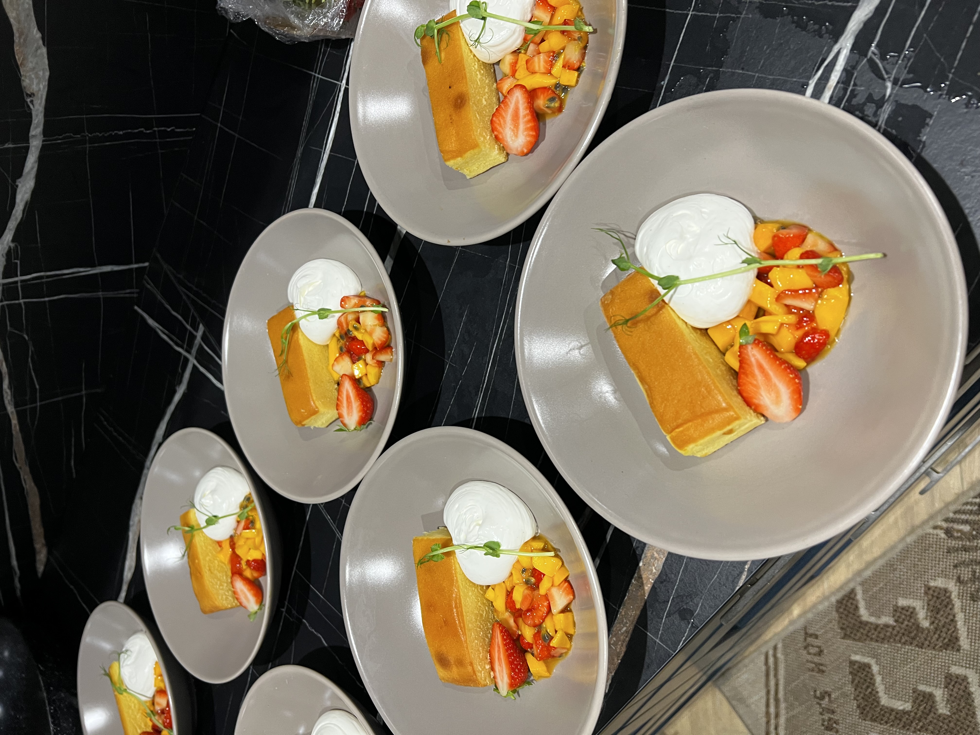 Multiple bowls each with a slice of sponge cake, a dollop of whipped cream, fresh strawberries, diced mango, and a green garnish on a black marble countertop.