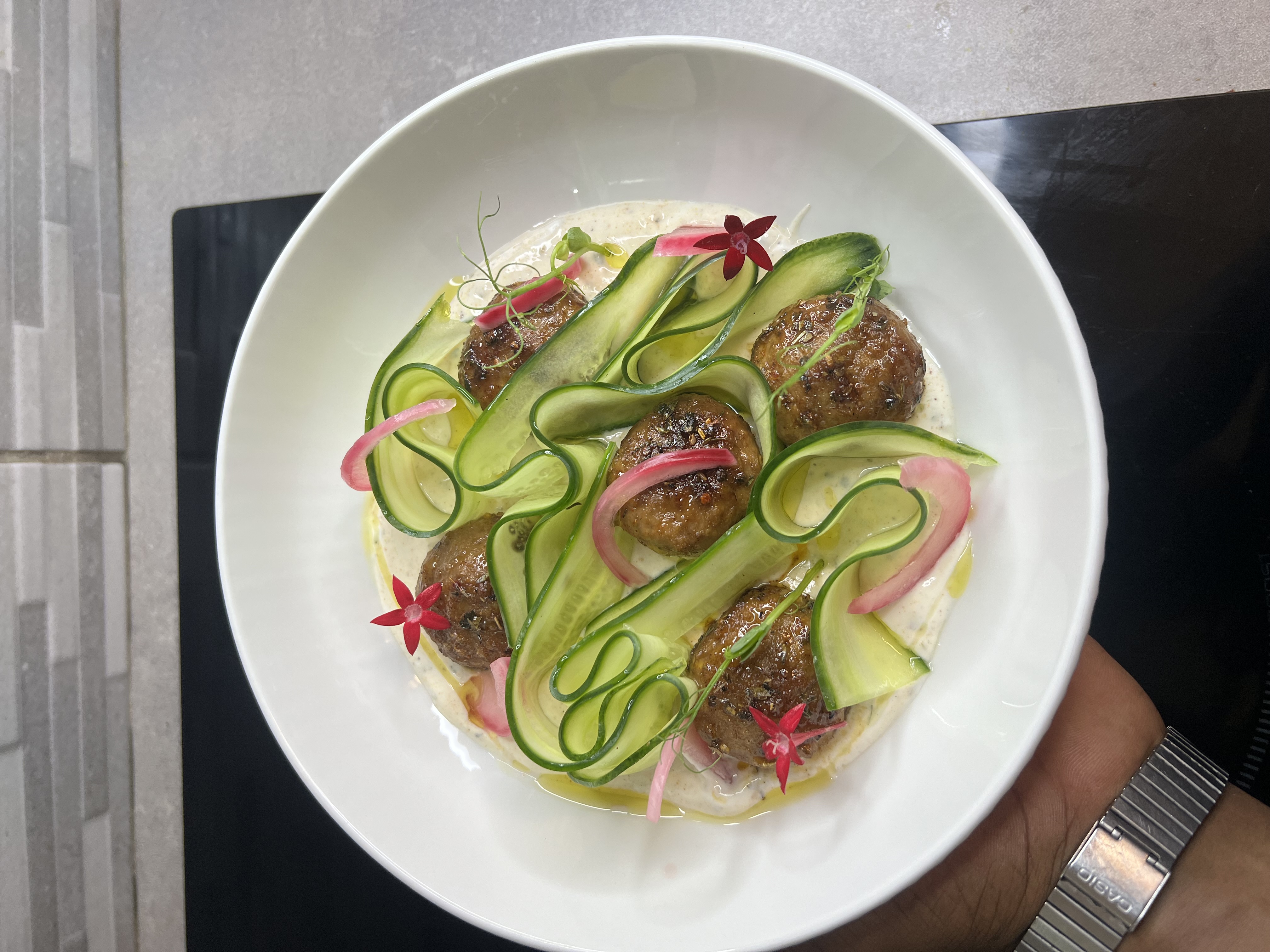 White bowl with grilled meatballs, thin cucumber ribbons, pickled red onions, edible red flowers, and creamy sauce drizzle.