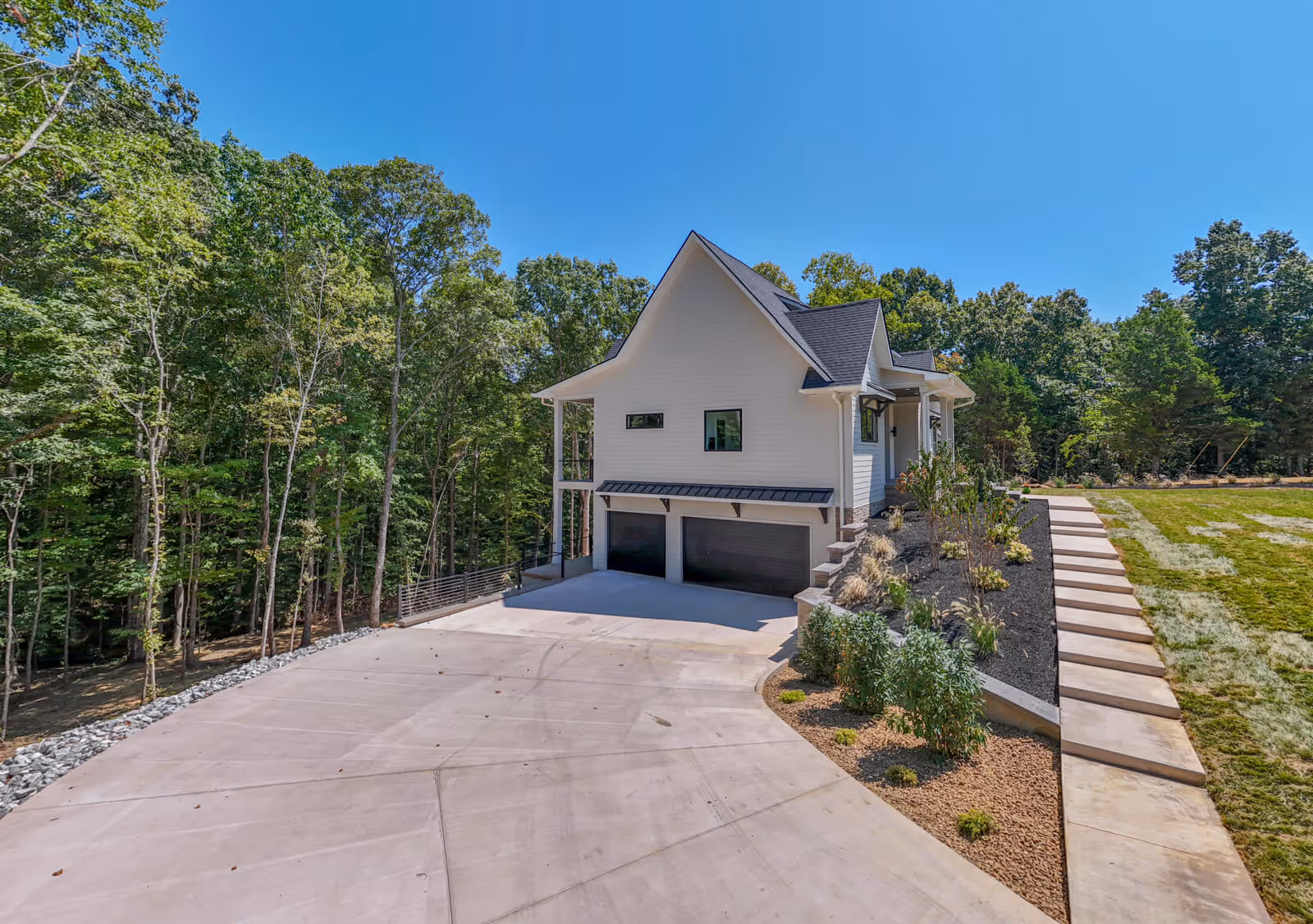 Modern white house with black roof, two-car garage, large concrete driveway, and landscaped garden beds, surrounded by trees under a clear blue sky.