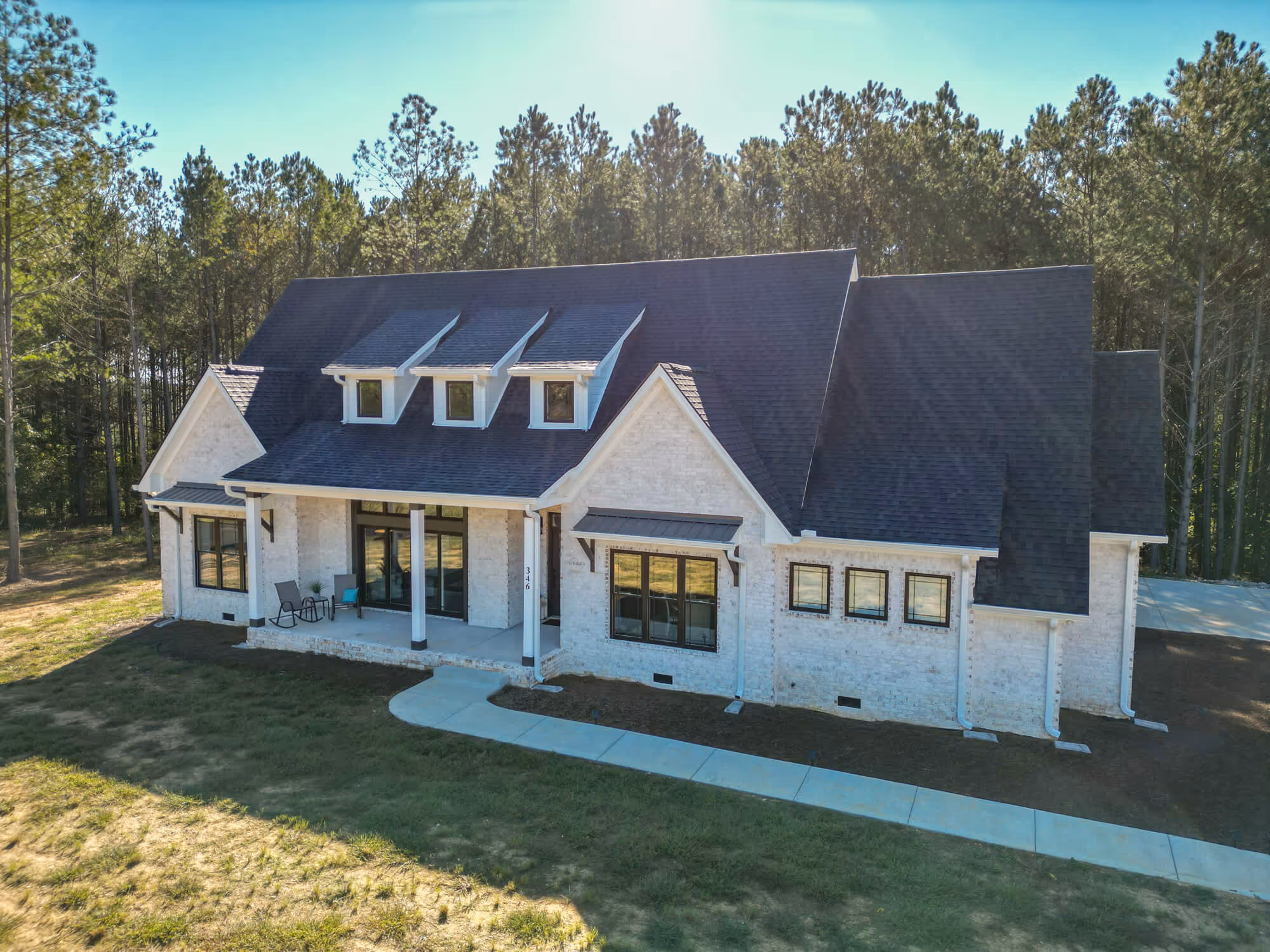 Modern white brick house with dark roof, three dormer windows, and a front porch set against a forest background.