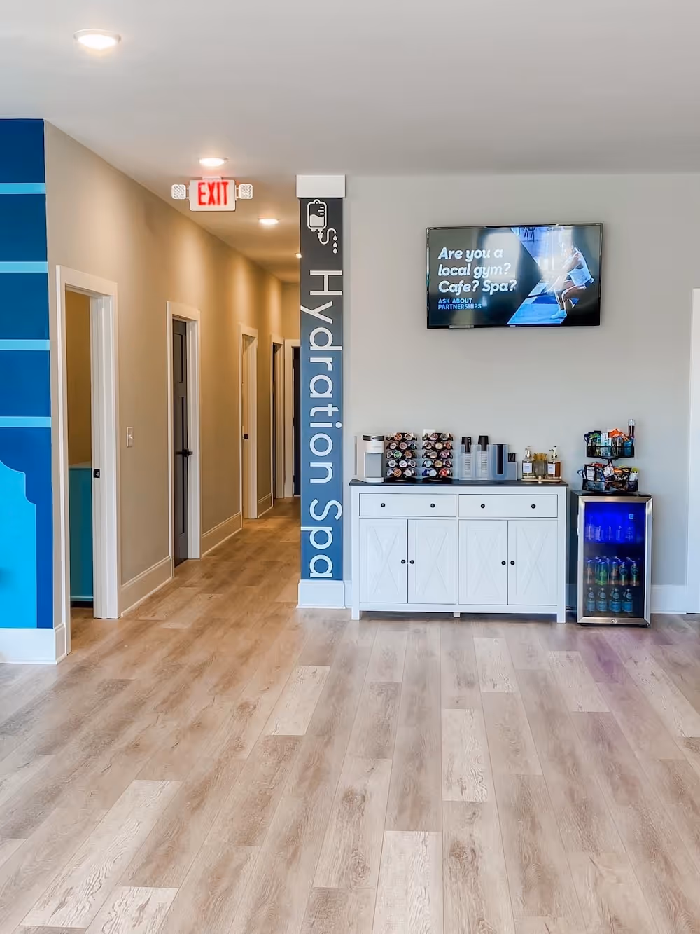 A modern hydration spa interior with light wood flooring, a sleek white cabinet stocked with wellness products, a small beverage fridge, and a mounted TV displaying information