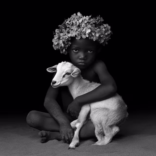 Black and white portrait of a child sitting on the ground, wearing a flower crown and hugging a young goat.