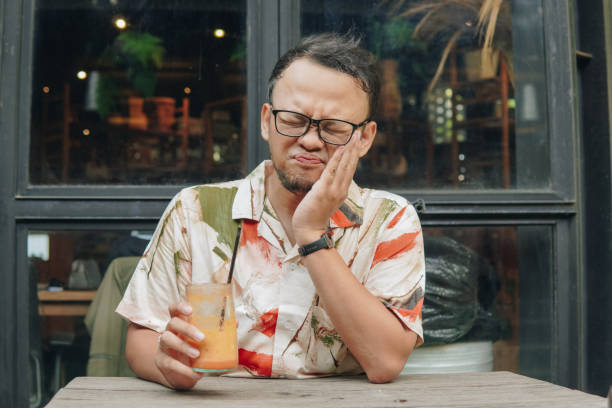 Sad and sick Asian man sipping his drink while holding his cheek, feeling pain or toothache. Sad and sick Asian man sipping his drink outside of a cafe while holding his cheek, feeling pain or toothache. TOOTHACHE SIPPING iced COFFEE stock pictures, royalty-free photos & images