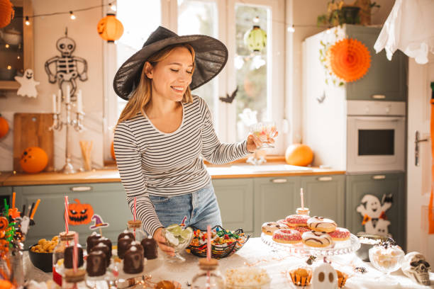 Woman preparing food table for Halloween Young woman in costume setting sweet table for Halloween lunch. A bowl of Halloween candy on a kitchen counter beside a woman holding her cheek in discomfort stock pictures, royalty-free photos & images