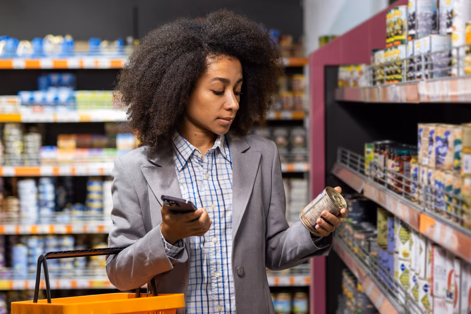 Woman in a gray blazer holding a can and a smartphone while shopping in a grocery store aisle.