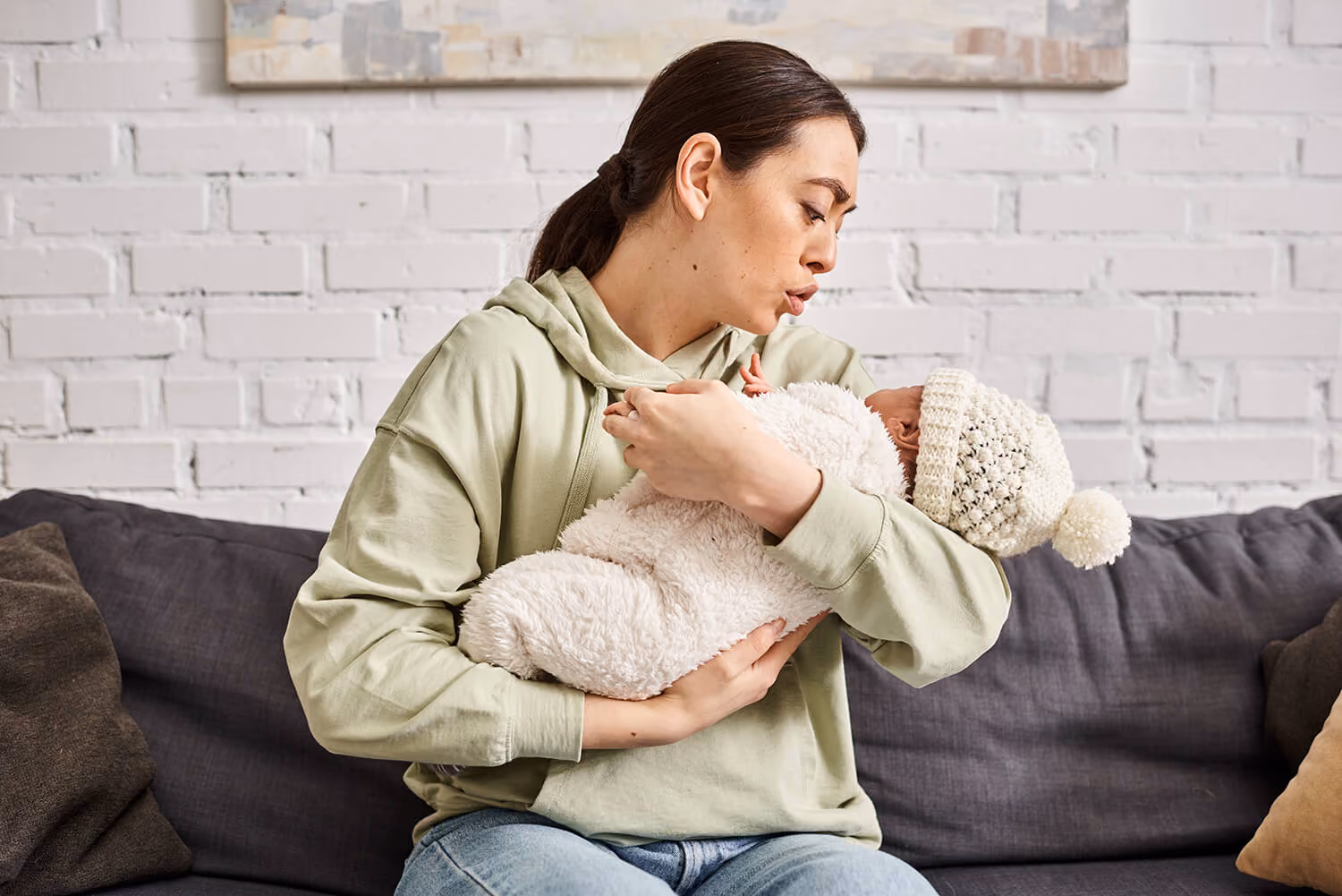 Woman in a light green hoodie sitting on a gray couch holding and looking lovingly at a baby wrapped in a fluffy white outfit and wearing a white knit hat with a pom-pom.