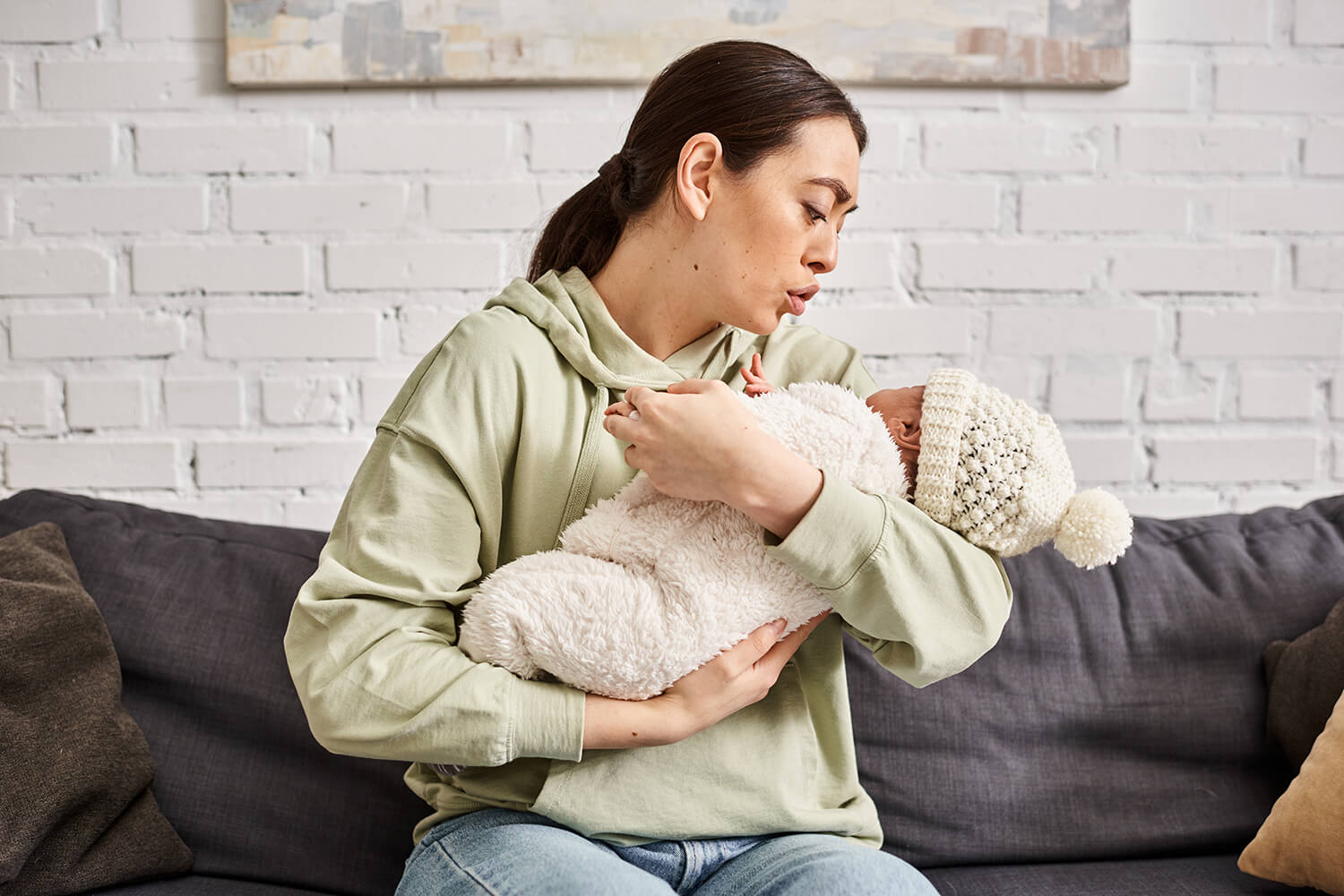 Woman in a light green hoodie sitting on a gray couch holding and looking lovingly at a baby wrapped in a fluffy white outfit and wearing a white knit hat with a pom-pom.