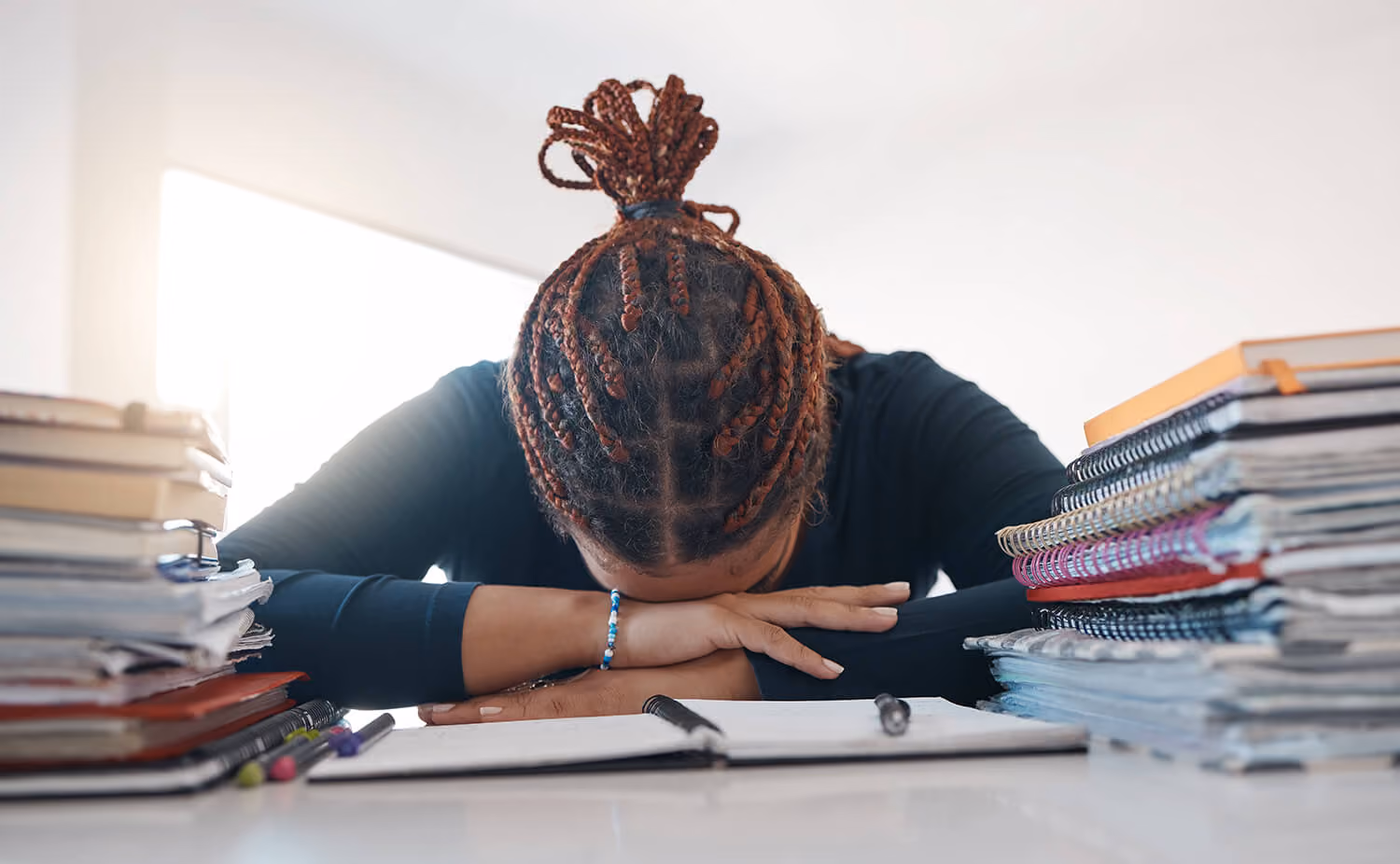 Person with braided hair resting head on crossed arms on a desk surrounded by stacks of notebooks and papers.