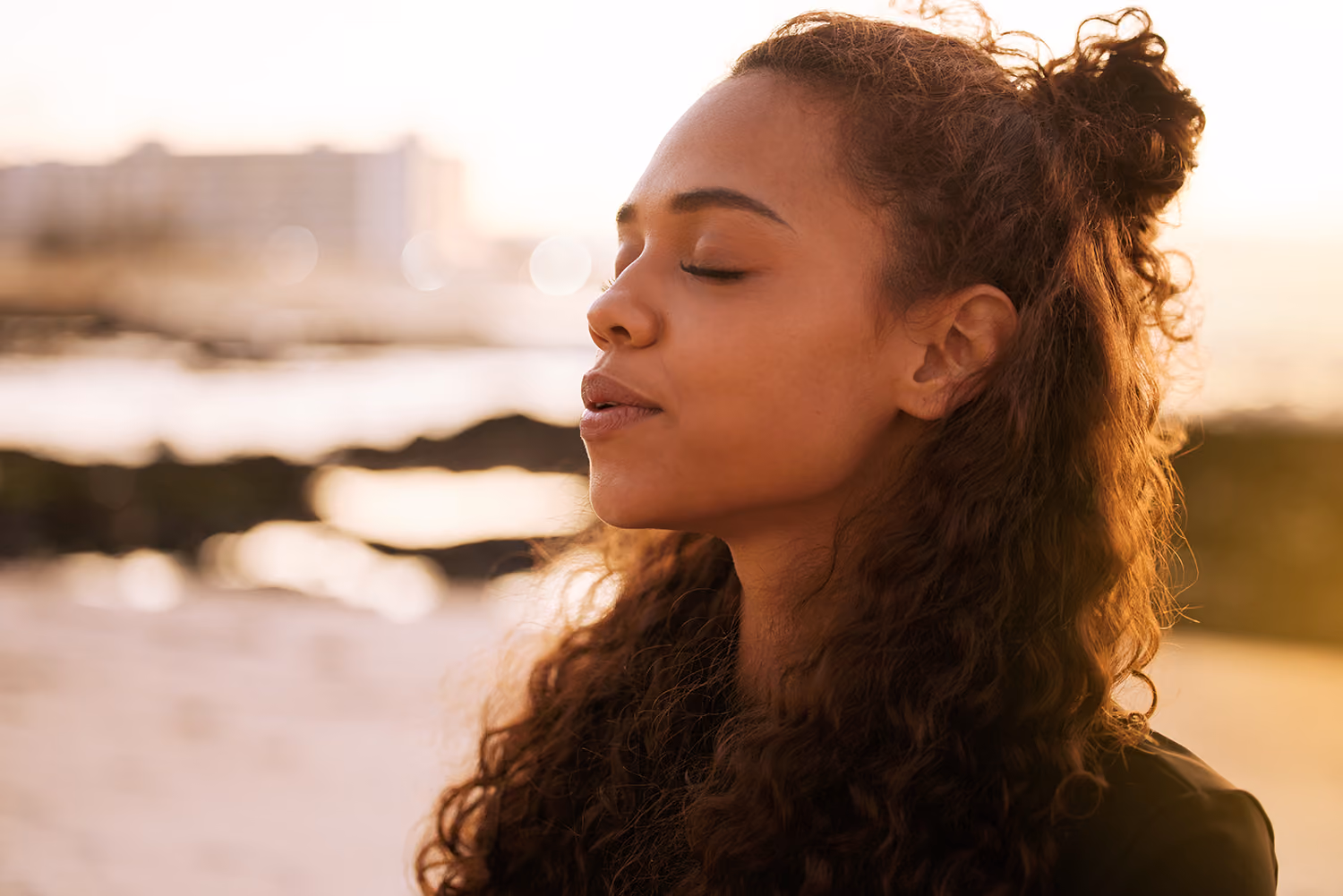 Young woman with curly hair tied back, eyes closed, standing outdoors near a body of water during sunset.