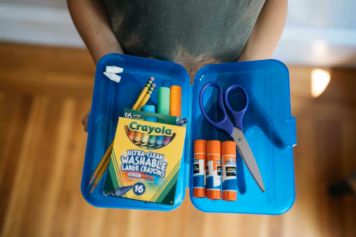 Child holding blue organizer filled with back-to-school supplies including crayons, pencils, glue sticks, and scissors