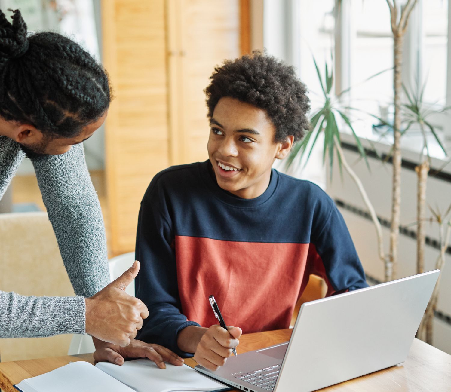 young man working at laptop and looking at his parent who is leaning towards him and giving him a thumbs up