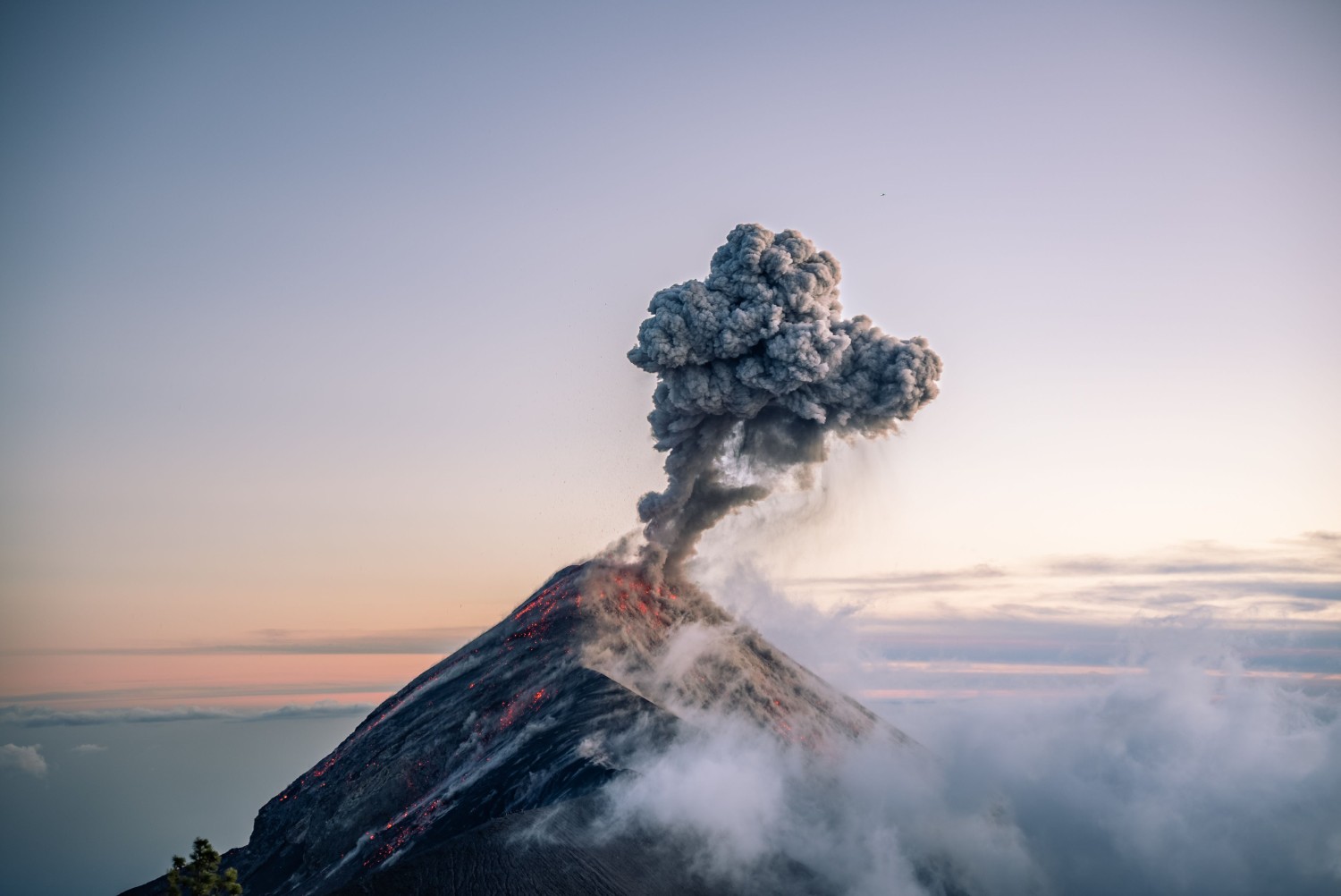 A volcano erupts at sunrise, sending a dark plume of ash into the sky as glowing lava and smoke spill down its slopes, surrounded by mist and clouds.
