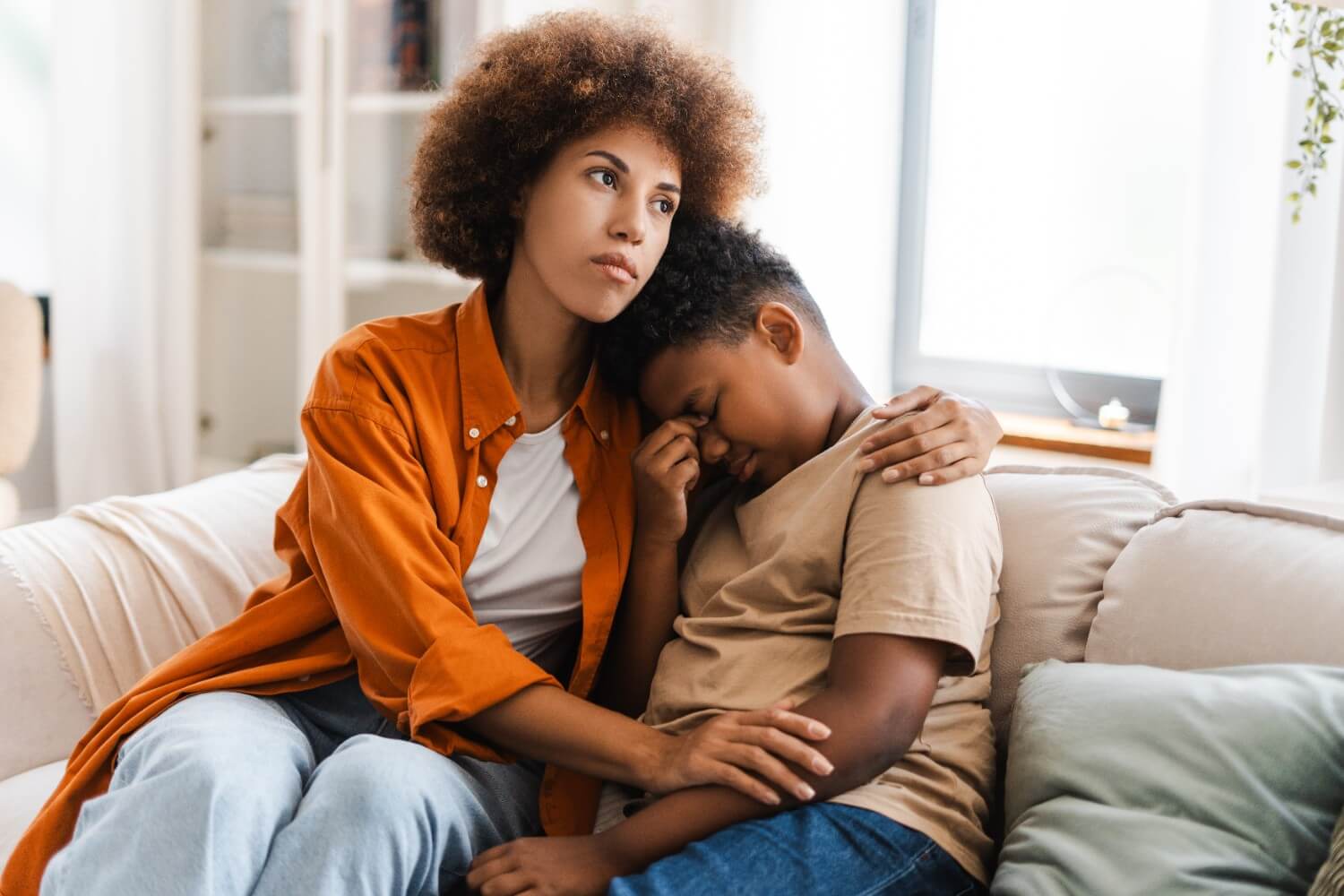 An adult woman sits on a couch, offering comfort to a young boy who is leaning his head on her shoulder with a distressed expression.
