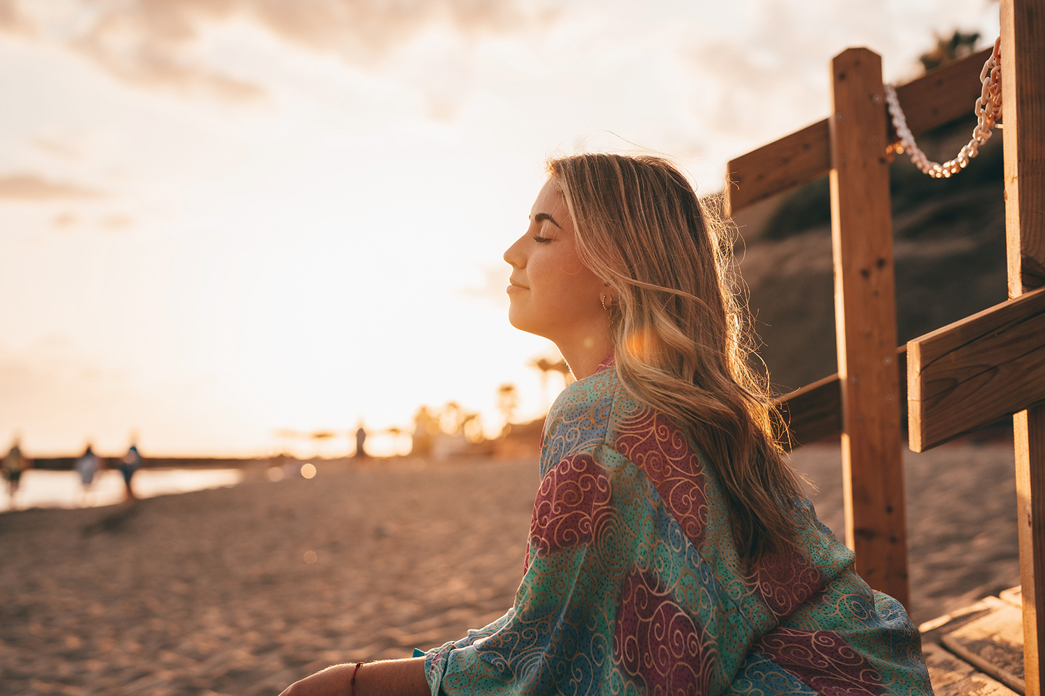 a young woman sits with her eyes closed on a sandy beach during sunset, enjoying a peaceful moment next to a wooden railing.