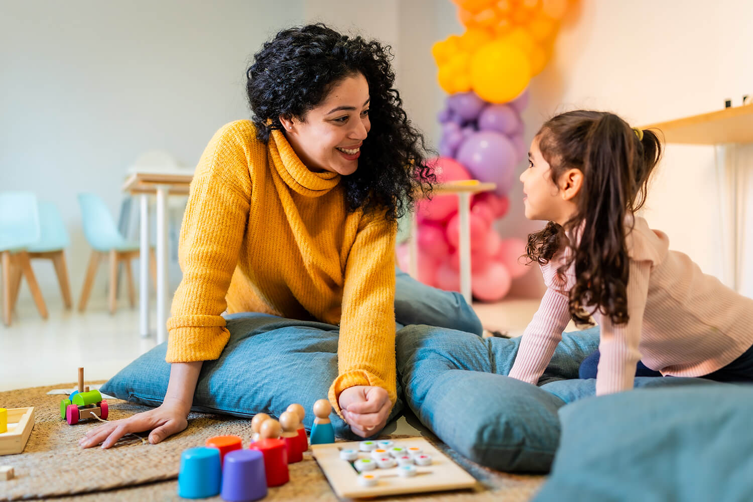 A smiling woman in a yellow sweater sits on floor cushions with a young girl while playing with colorful wooden toys in a bright room.
