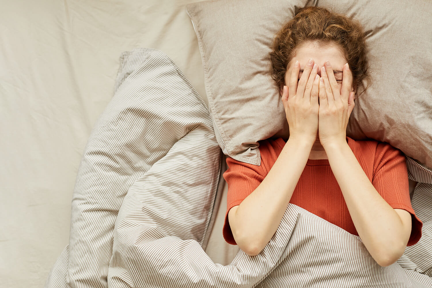 An overhead view of a woman lying in bed covering her face with her hands, looking stressed or anxious.