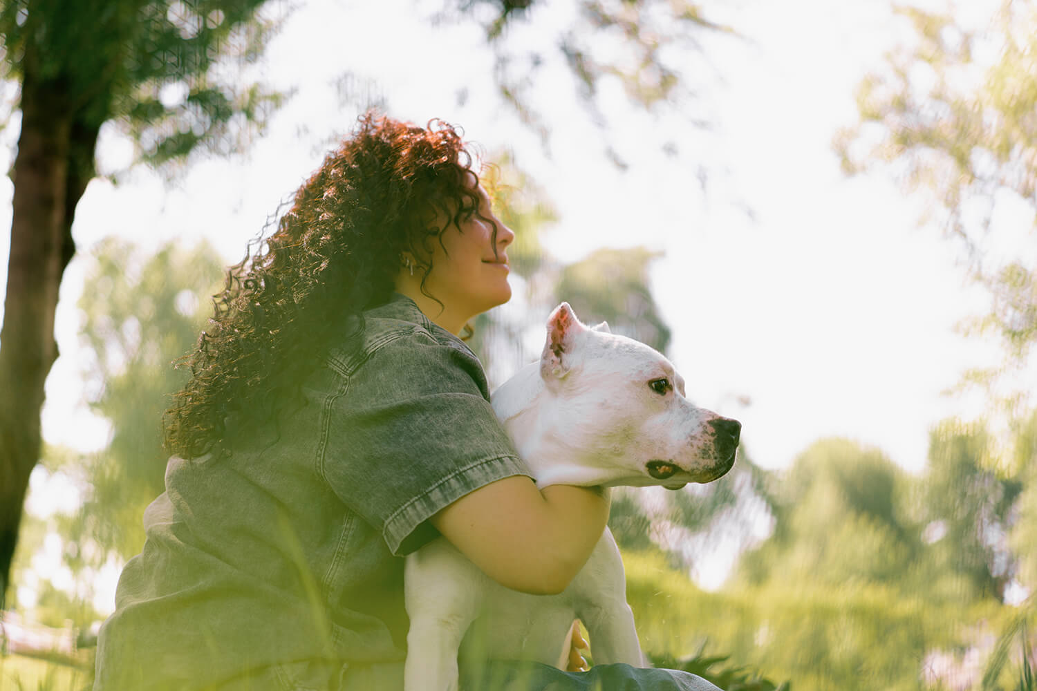 A smiling woman with dark curly hair hugging her white dog outdoors in warm daylight, illustrating a peaceful moment of connection.