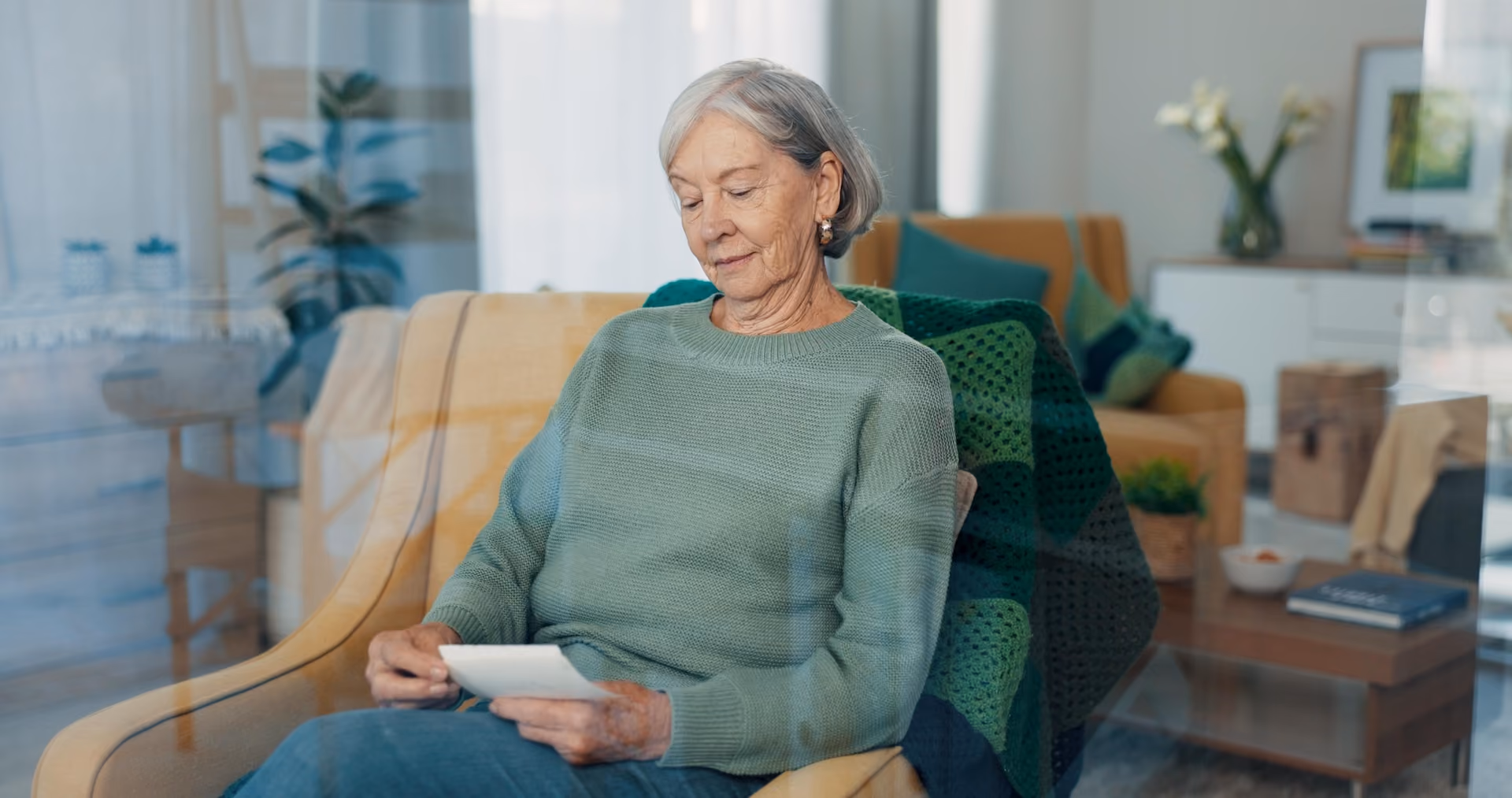 Elderly woman in a green sweater sitting in a cozy living room chair, looking at a photo.