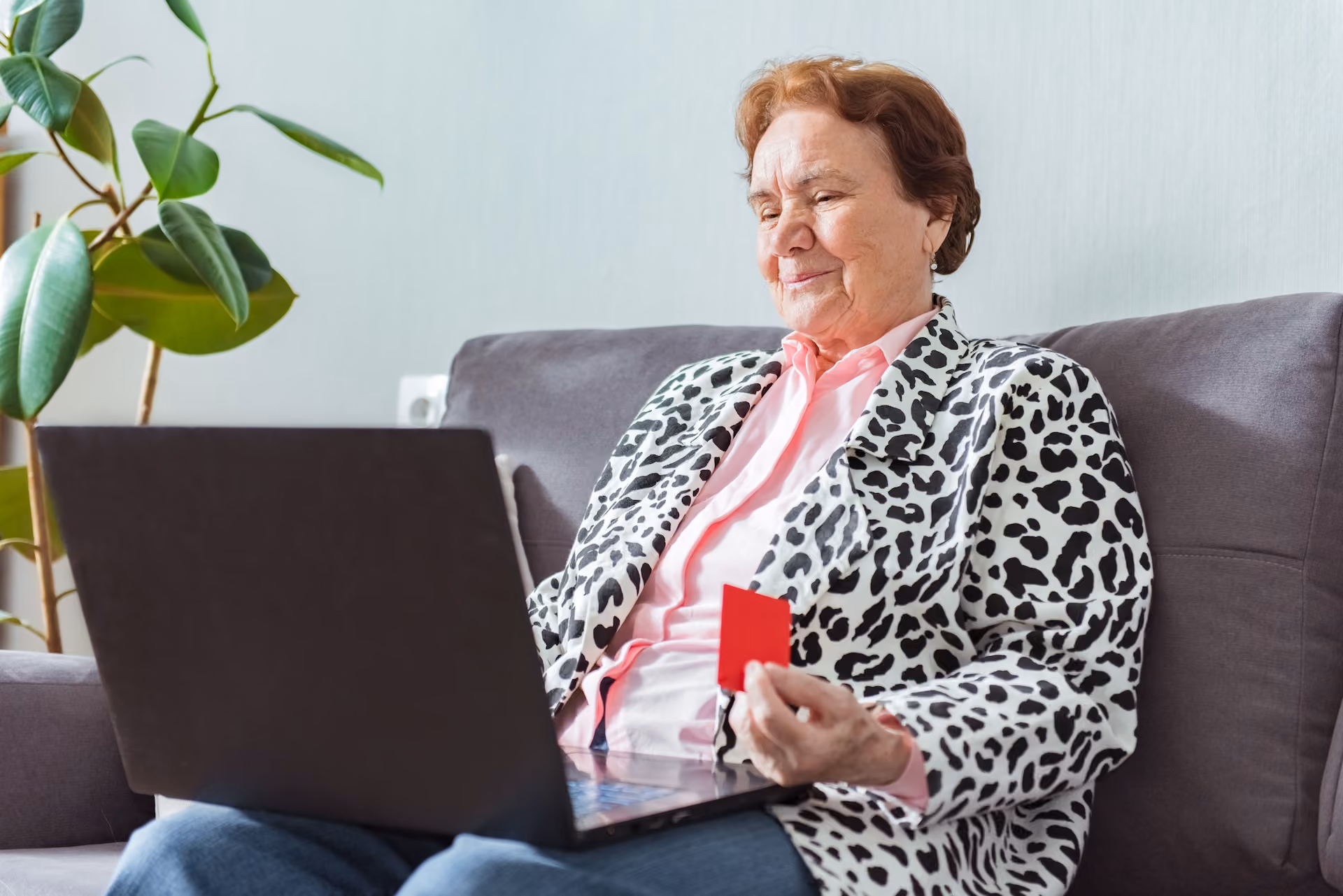 Elderly woman sitting on a gray couch holding a red card while using a laptop.