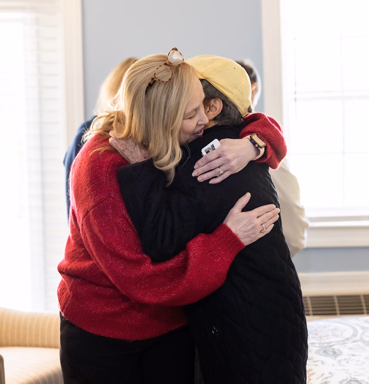Two women embracing warmly indoors, one wearing a red sweater and glasses on her head, the other in a yellow cap.