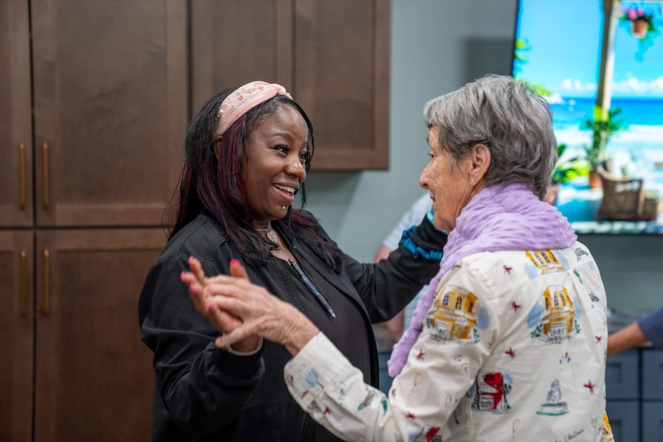 Smiling woman warmly holding hands and engaging with an elderly woman in a cozy indoor setting.
