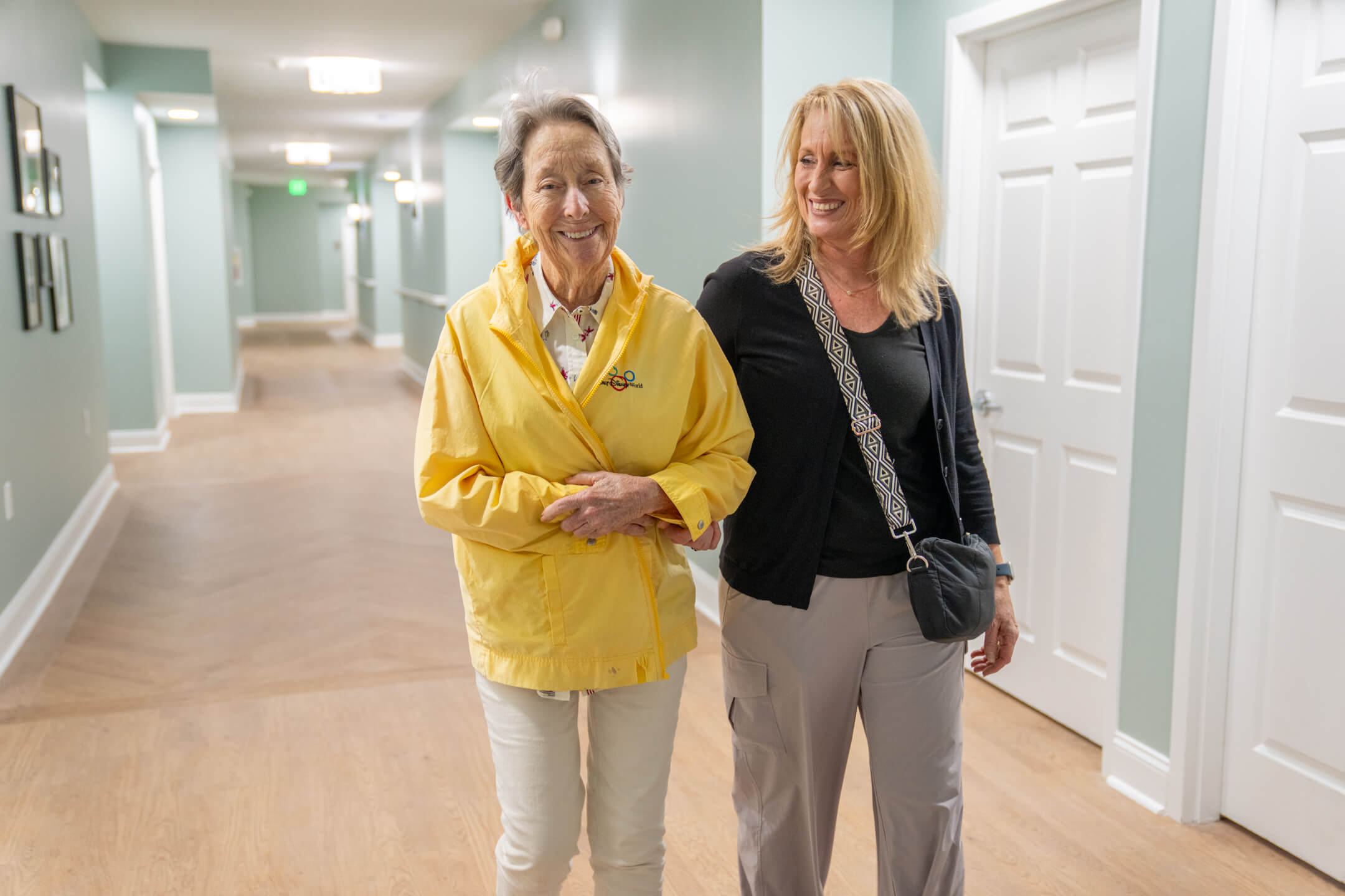 Smiling elderly woman in yellow jacket walks arm-in-arm with middle-aged woman in hallway.