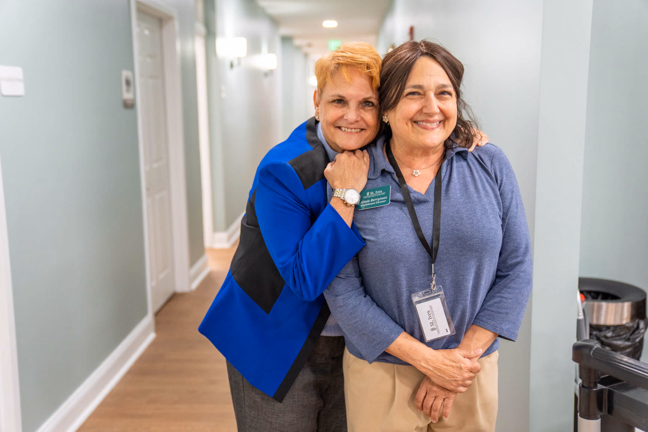 Two smiling women in a hallway, one hugging the other from behind with their arms around her shoulders.