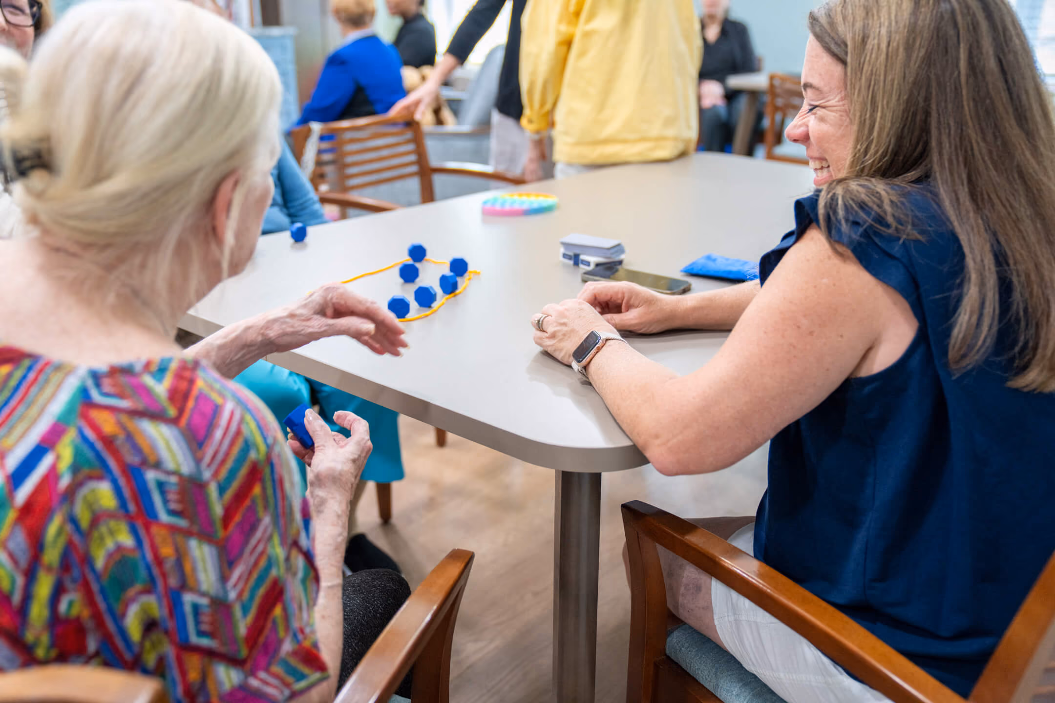 Two women sitting at a table playing with blue hexagonal blocks and a yellow string.