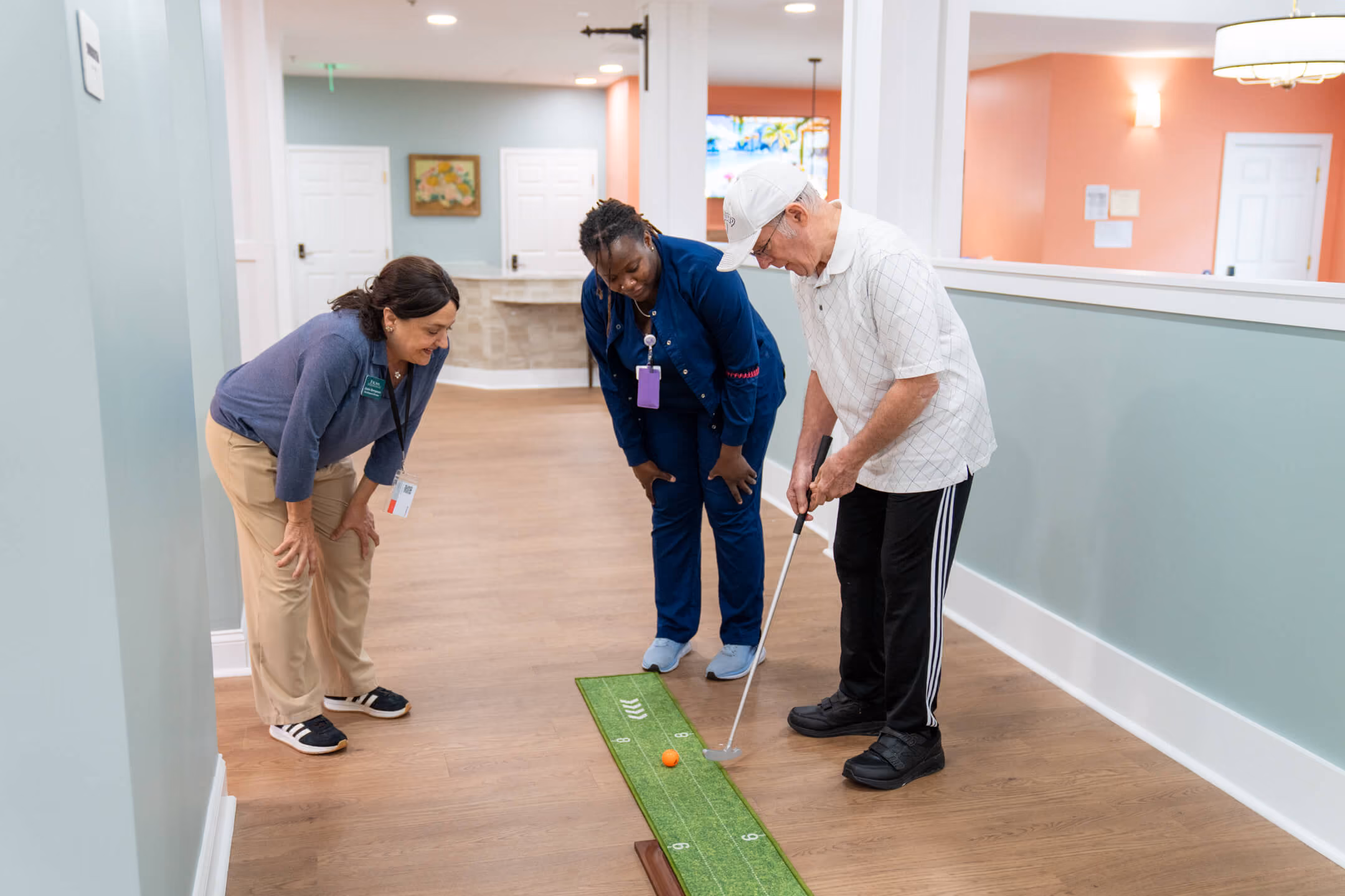 Elderly man putting golf ball on indoor putting mat with two women watching and smiling.