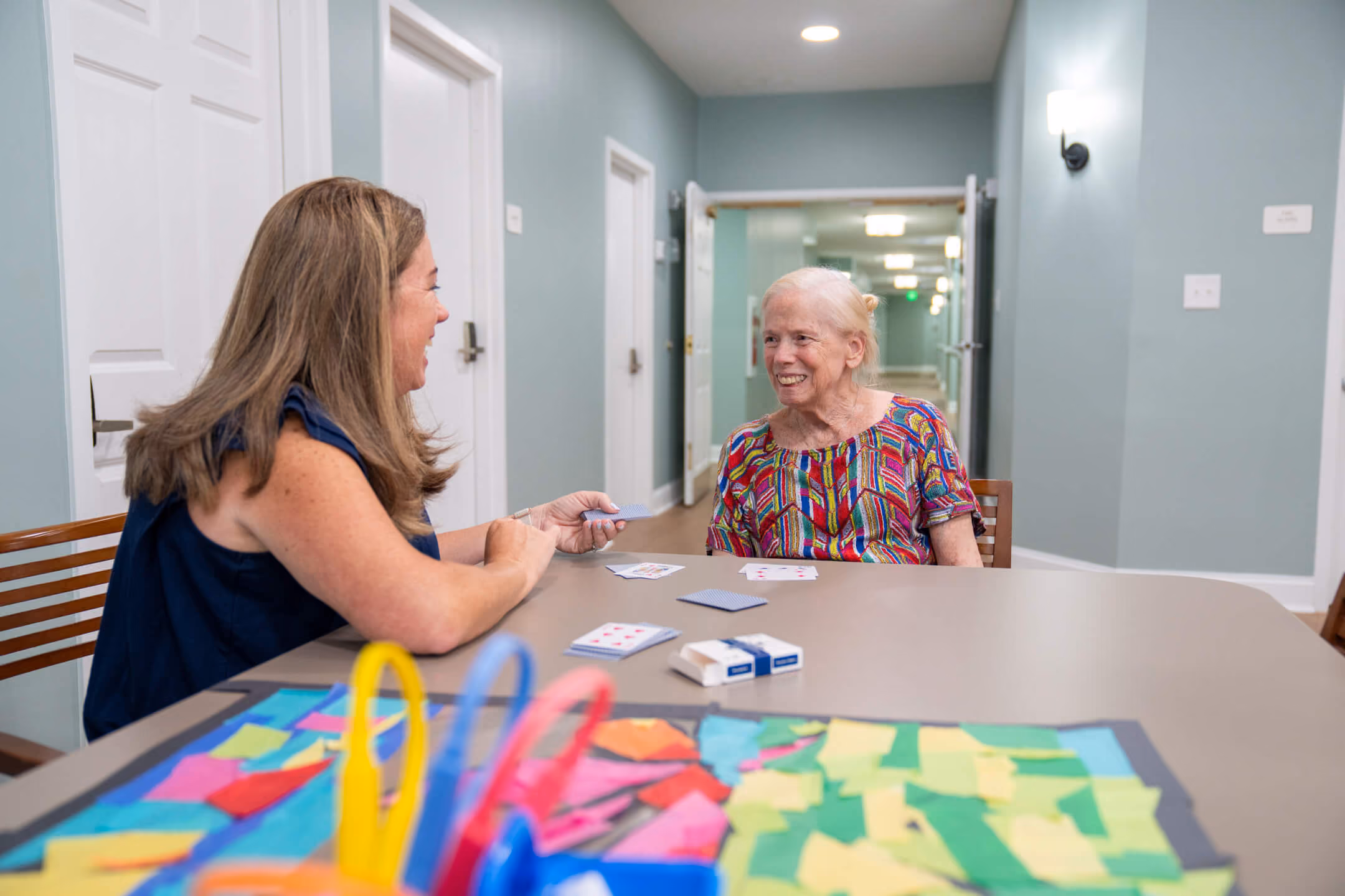 Two women smiling and playing cards at a table in a brightly lit room.