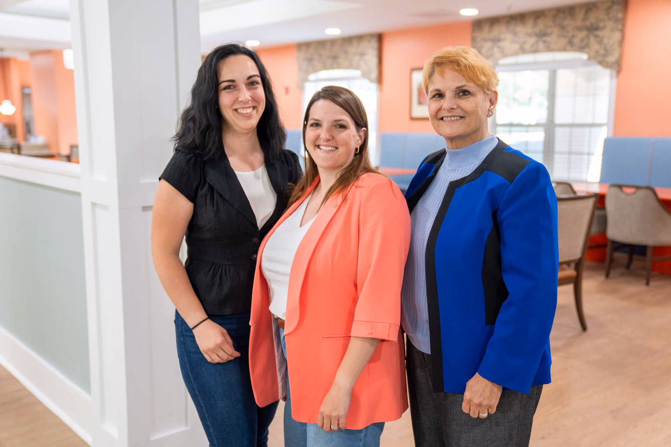 Three smiling women standing indoors in casual and business casual clothes.