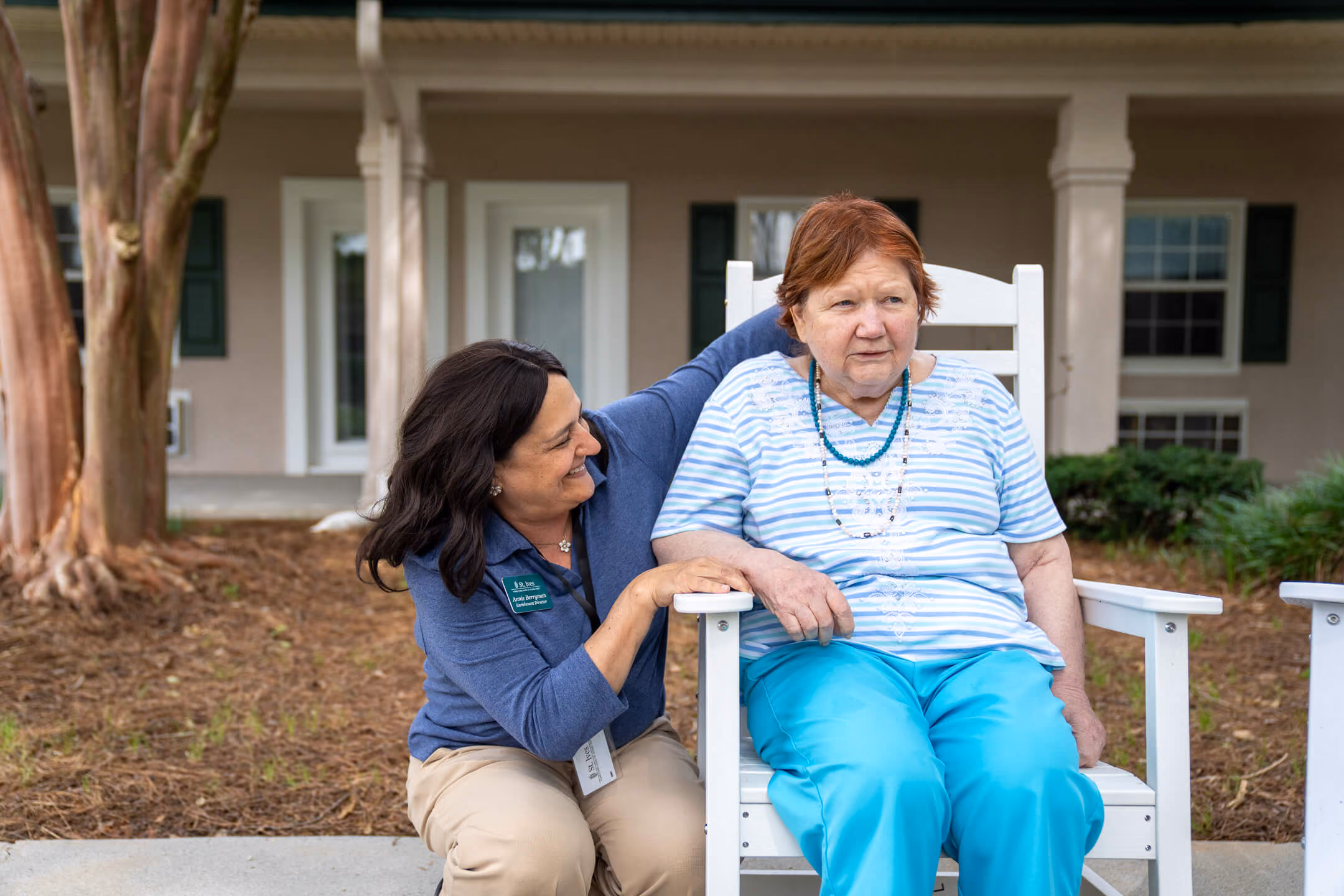 Caregiver smiling and leaning toward elderly woman sitting on a white wooden chair outside a building.