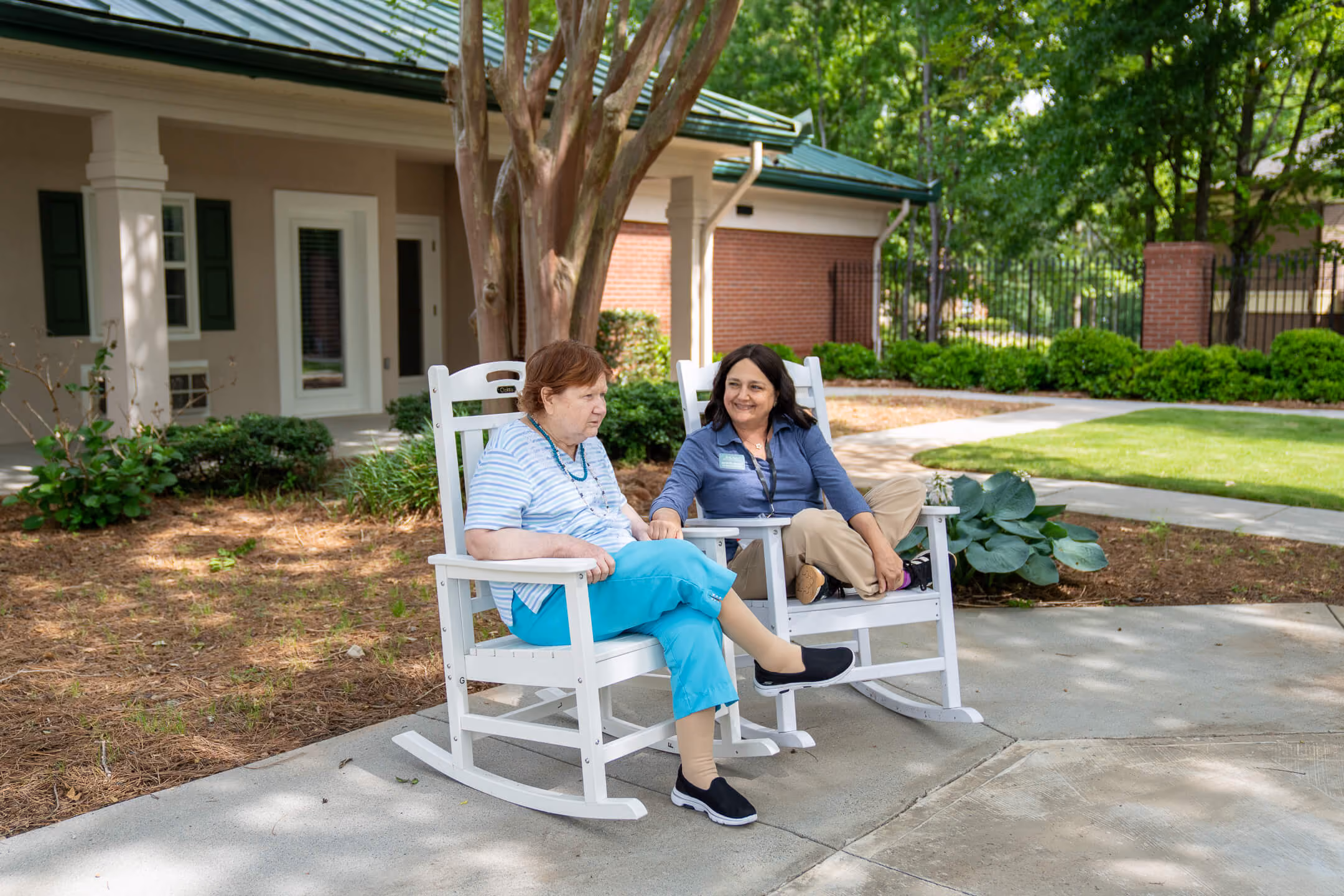 Two women sitting and talking on white rocking chairs outside a building with garden greenery.