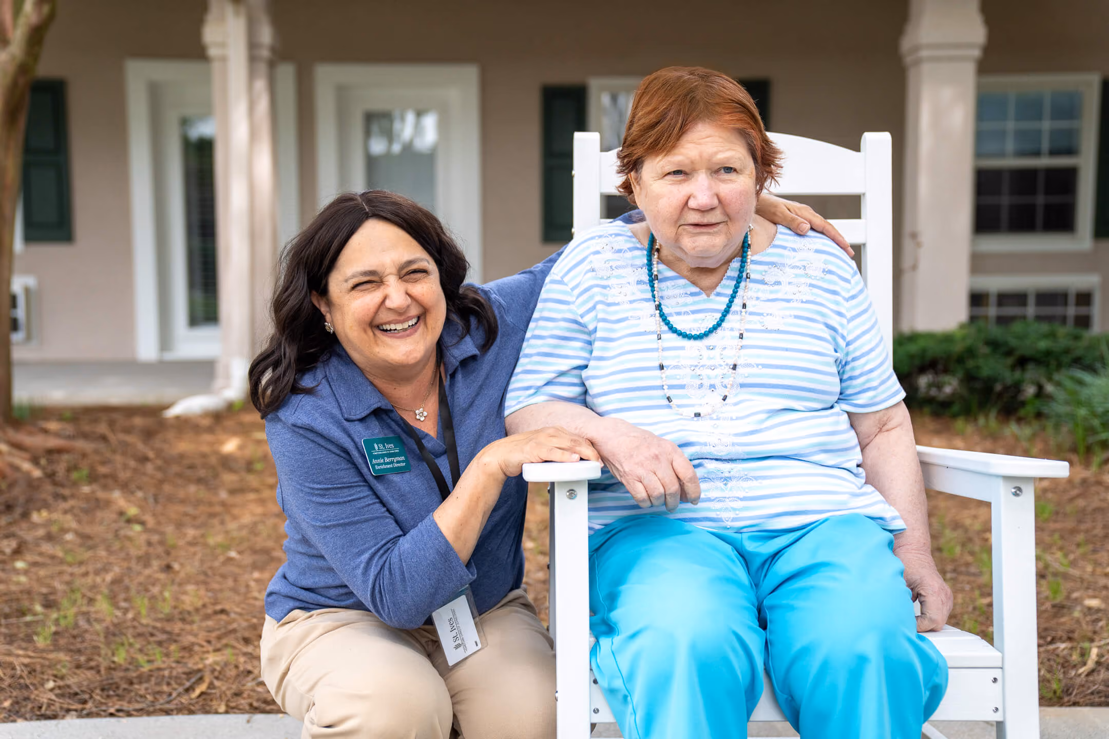 Smiling woman crouching beside an elderly woman seated in a white rocking chair outside a residential building.