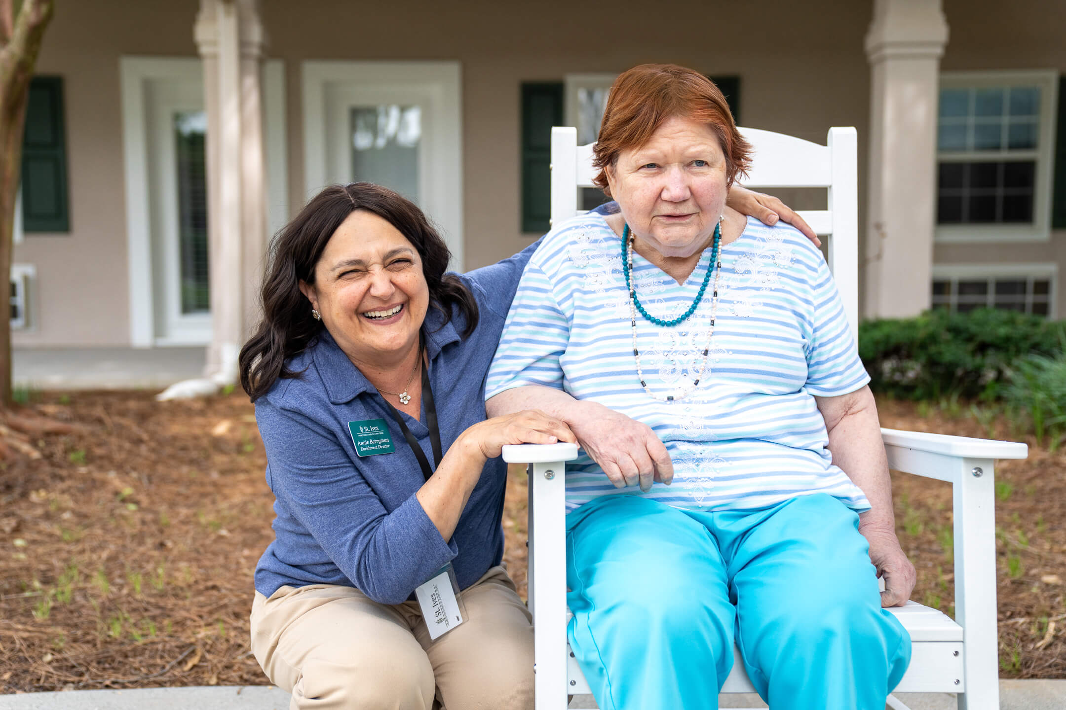 Smiling woman crouching beside an elderly woman seated in a white rocking chair outside a residential building.