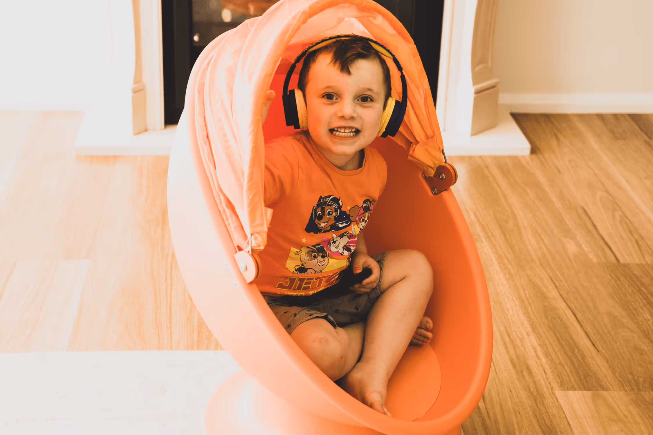 A young boy sits on an orange chair and wears yellow and black headphones.