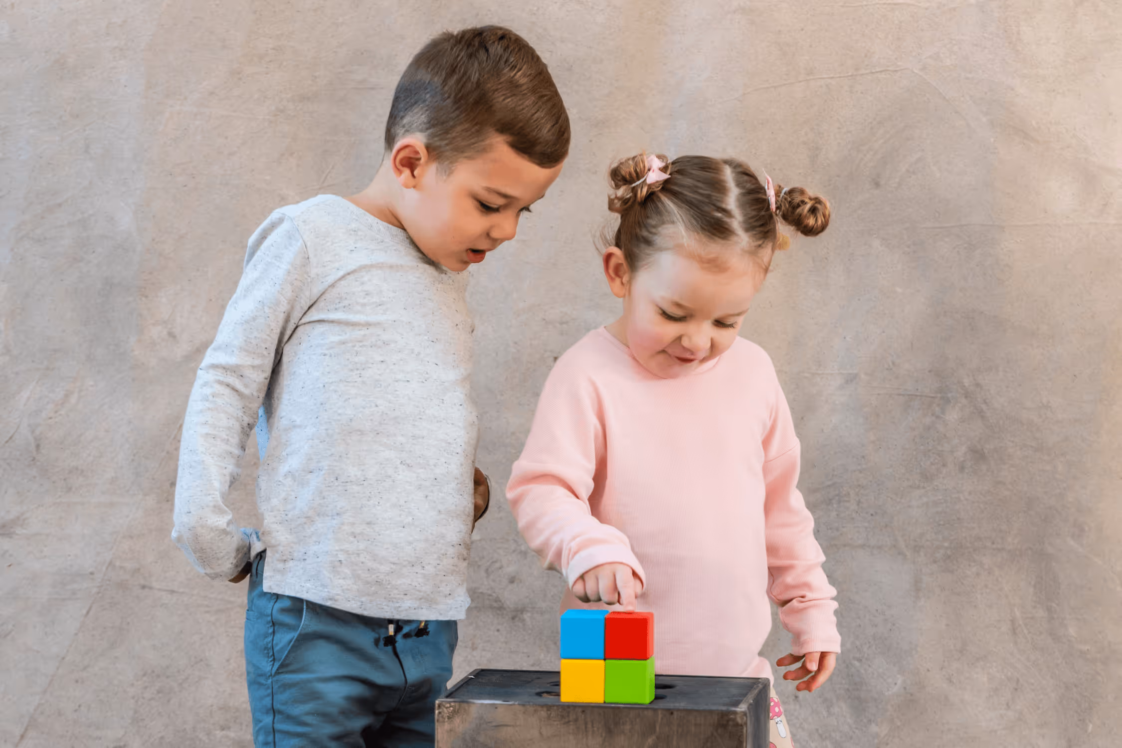 A young autistic boy wearing a grey shirt stands next to a younger girl with a pink jumper. They are both staring at a stack of coloured blocks on a wooden bench and the girl is pointing at them.