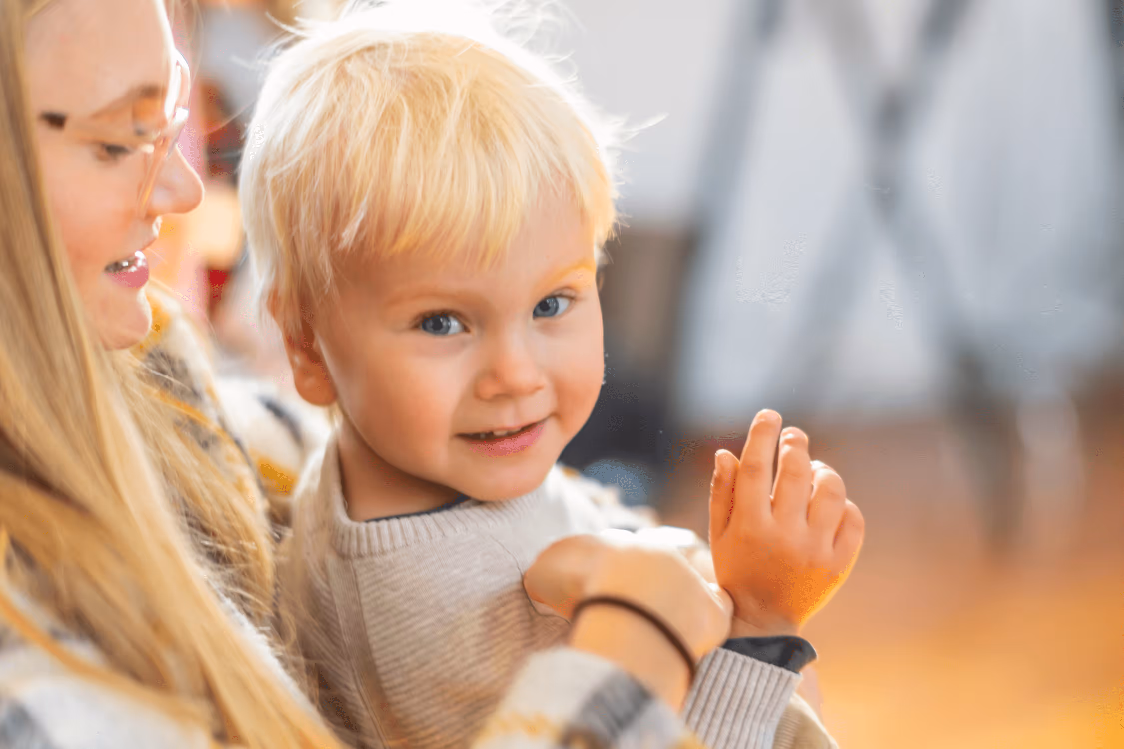 A young autistic boy with blonde hair and blue eyes sits on his mother's lap and smiles.