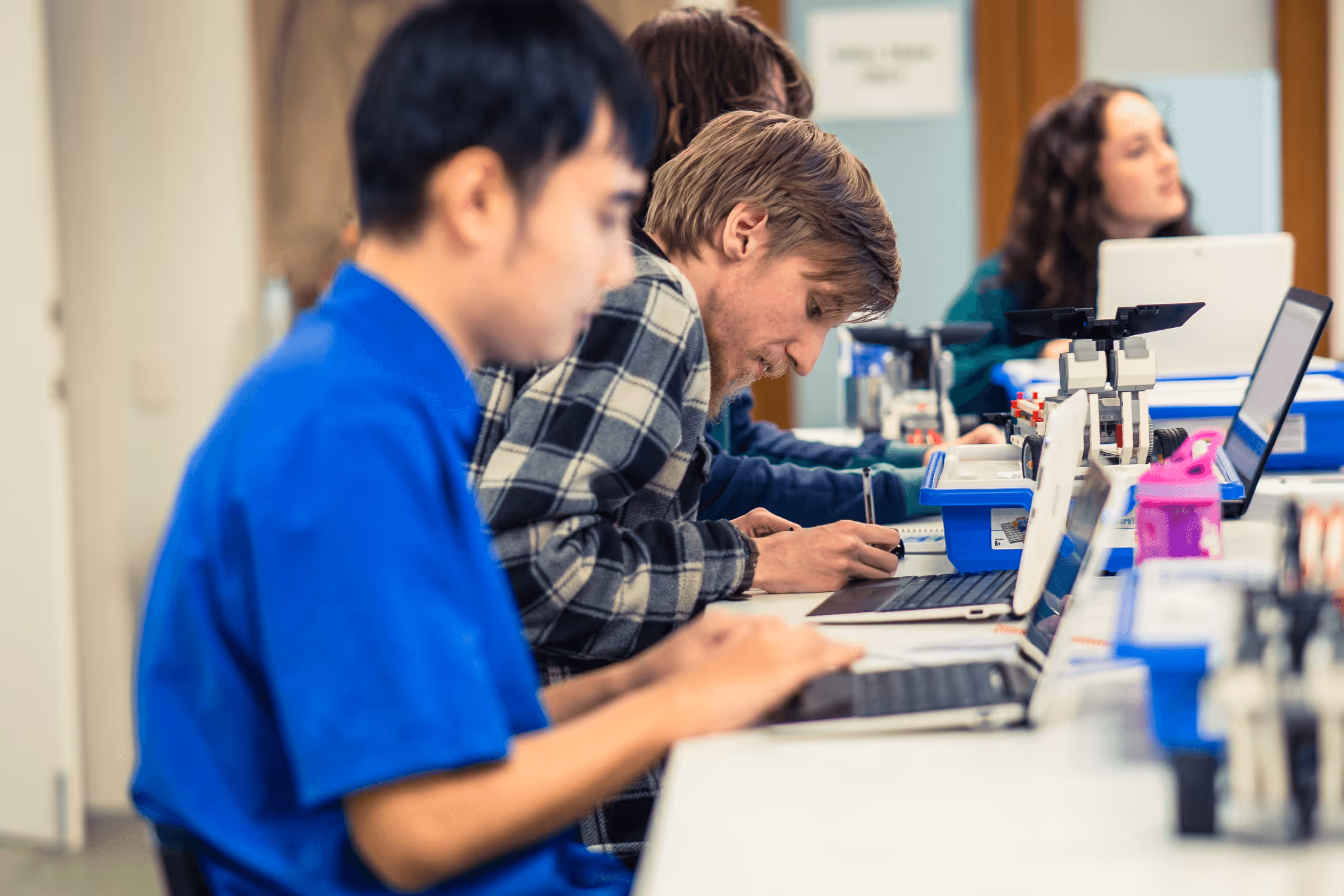 A young autistic man sitting at a desk with other young people writing with a black pen.