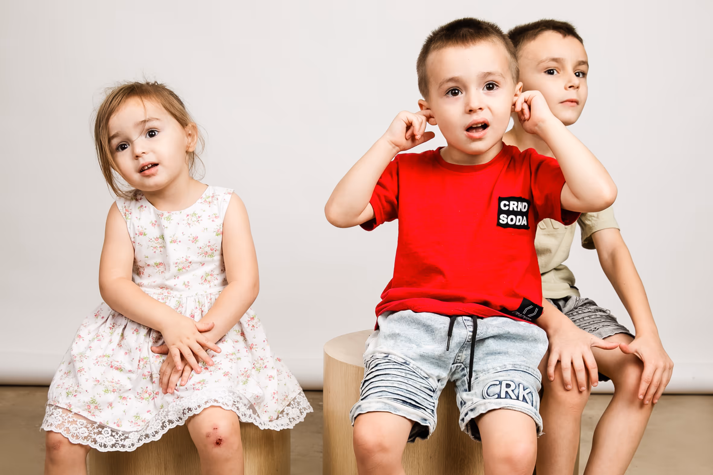 Three young children with brown hair sitting on wooden blocks in front of a plain grey background.