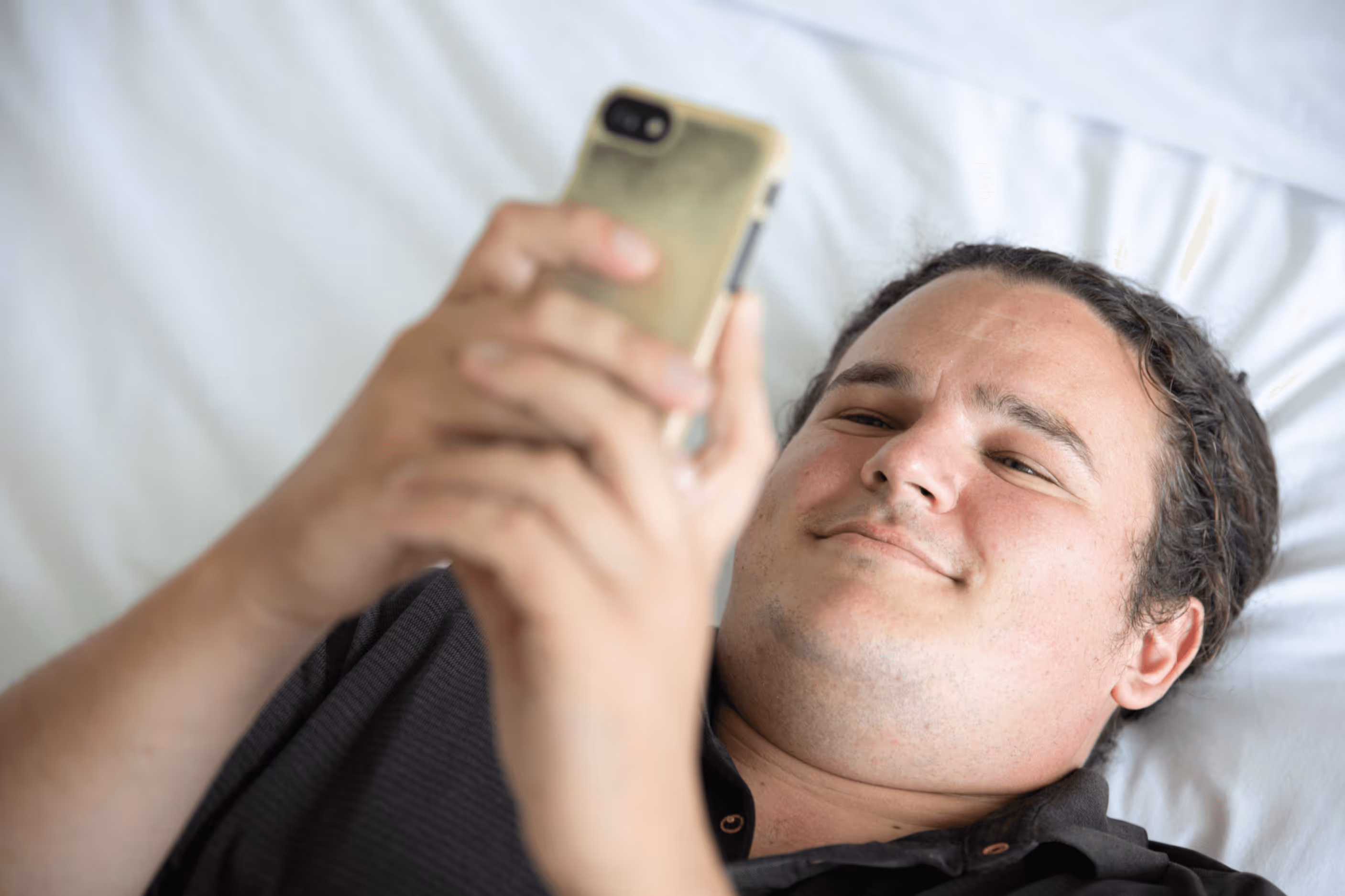 An autistic man with a black shirt lying on a bed with white sheets and scrolling on a phone.