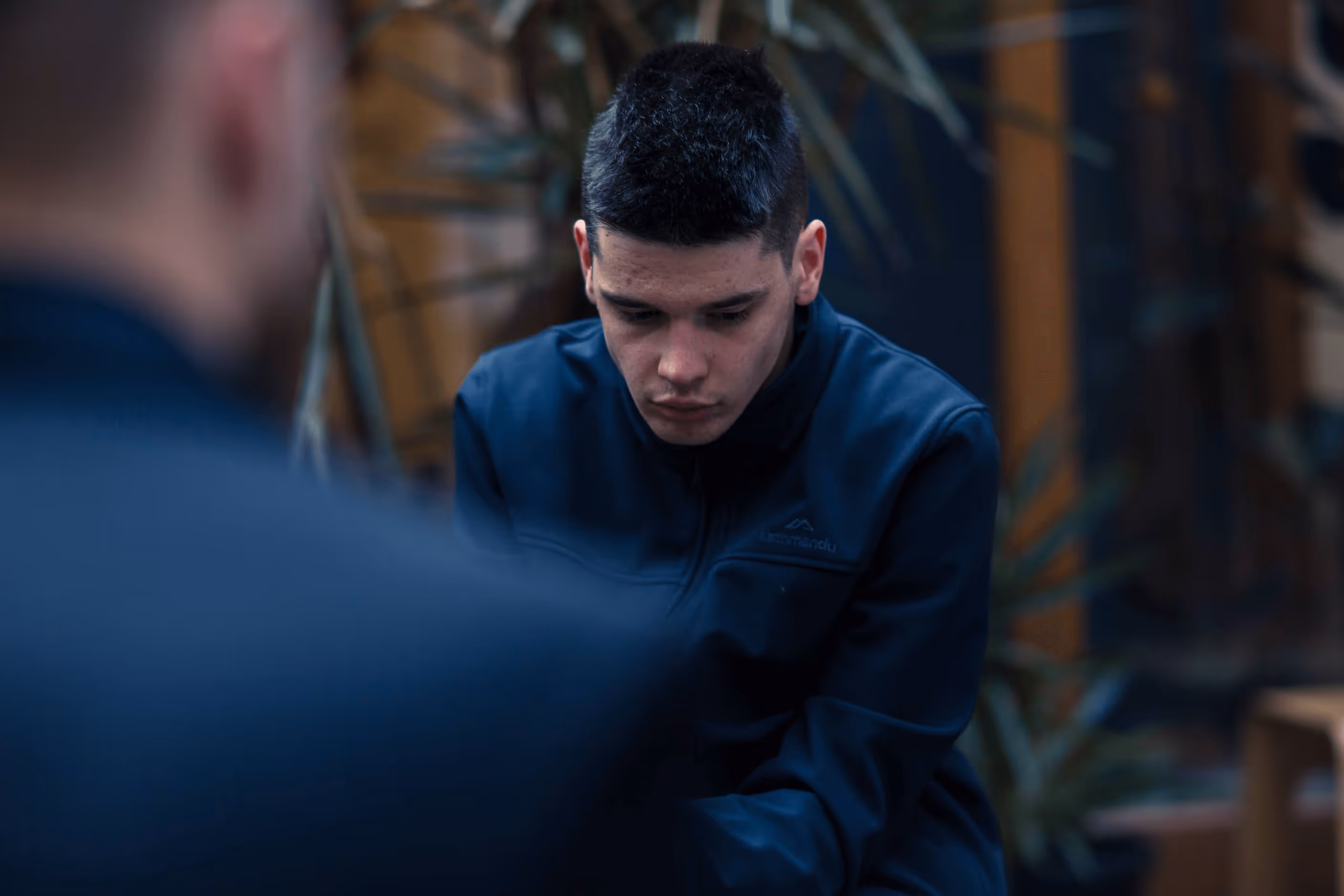 A young man with autism wearing a dark jacket sits and looks at the ground.