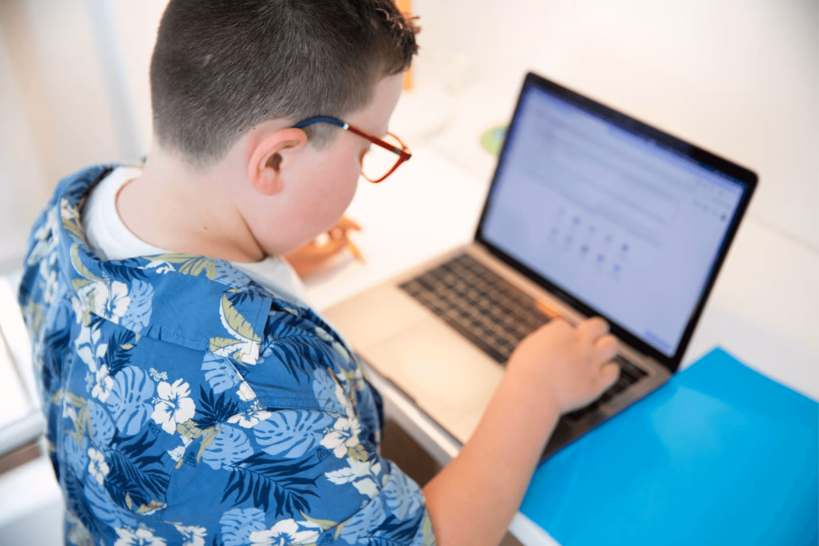 A boy with brown hair, red glasses and a blue shirt with palm trees on it typing on a silver laptop at a white desk.