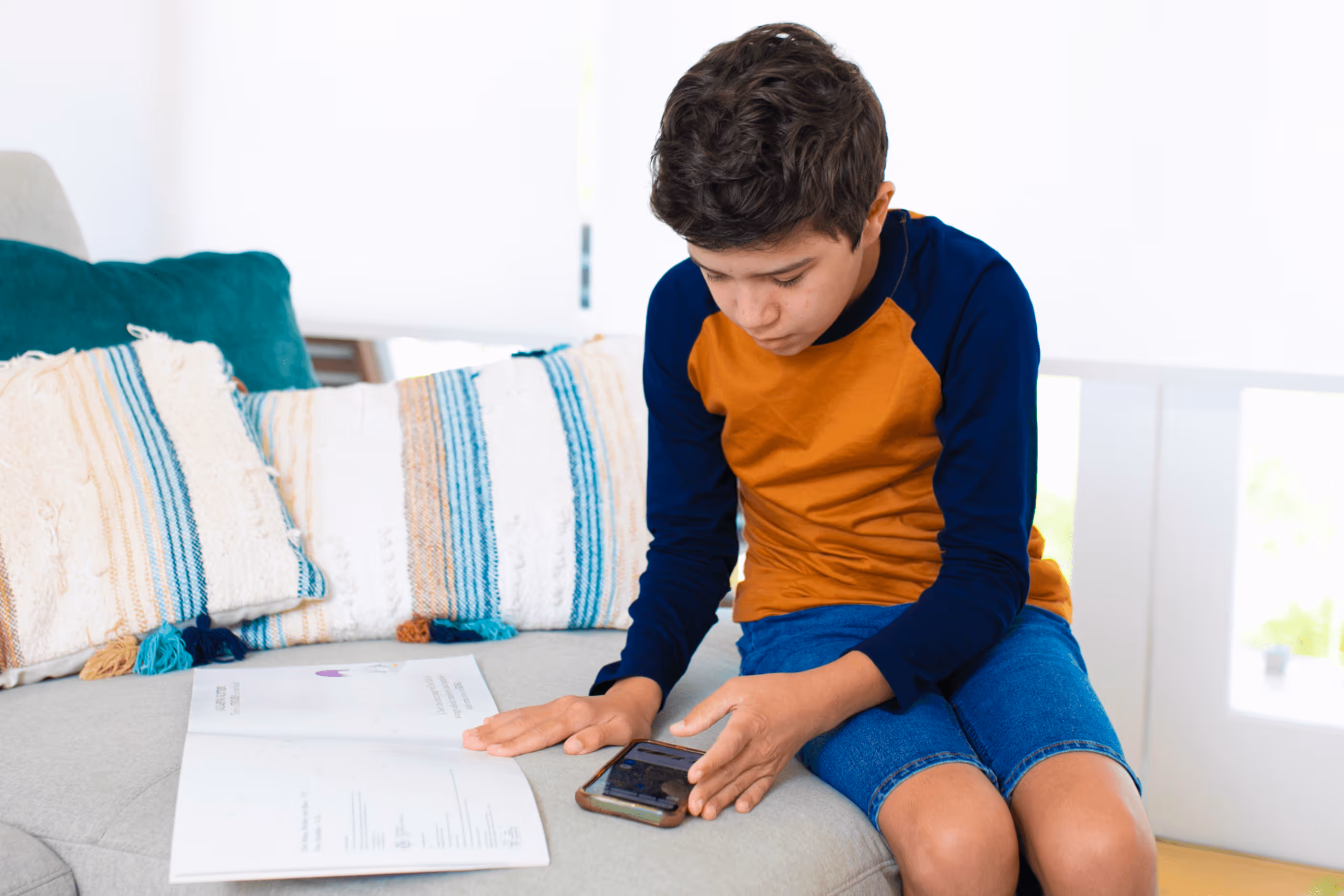 A boy with brown hair wearing a long-sleeve shirt and shorts sits on a grey couch and taps his finger on a mobile phone. Next to him is a picture book, which he holds open with his other hand.