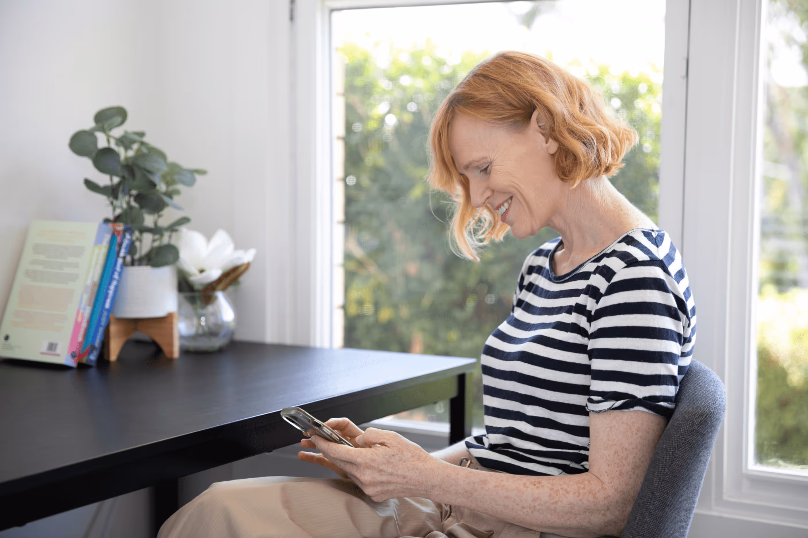 A woman with short red hair wearing a short-sleeve black and white shirt is using her phone and smiling at the screen. She is sitting in a grey chair at a black desk with a window in the background showing green plants outside.