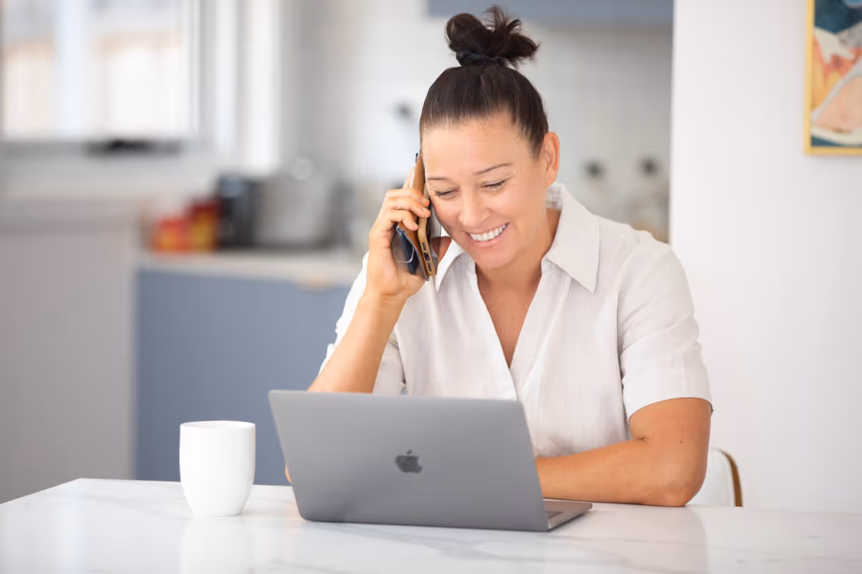 A mum with dark brown hair tied in a bun and a white collared shirt sits in front of a silver laptop and holds a phone with a tan case to her ear, smiling.