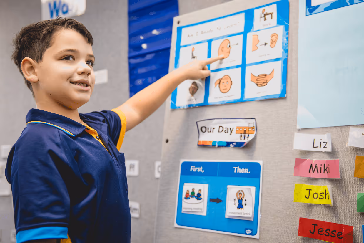 An autistic boy with short, dark hair points at a laminated visual support on a grey wall.