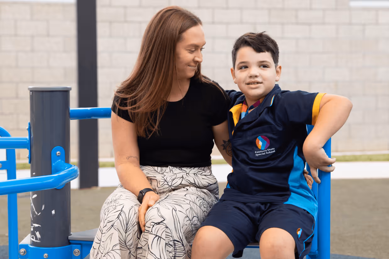 An autistic boy and a support teacher sitting on play equipment and smiling.