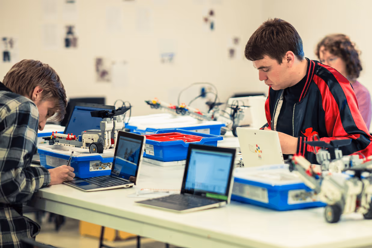 Two autistic men sit at a table in front of laptops at a large white desk.